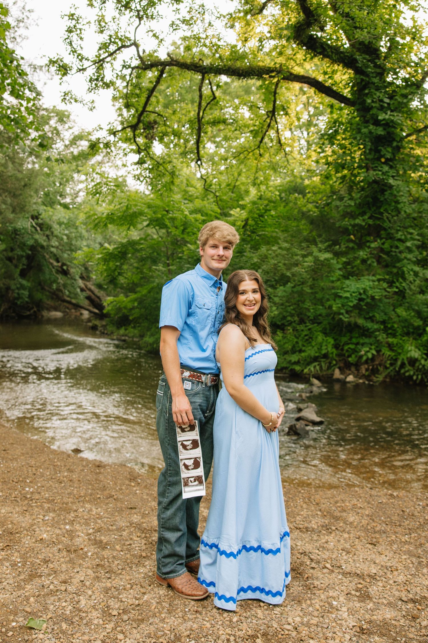 Pregnant couple poses by a creek, woman in blue maxi dress, man holding ultrasound photos, surrounded by lush green trees.