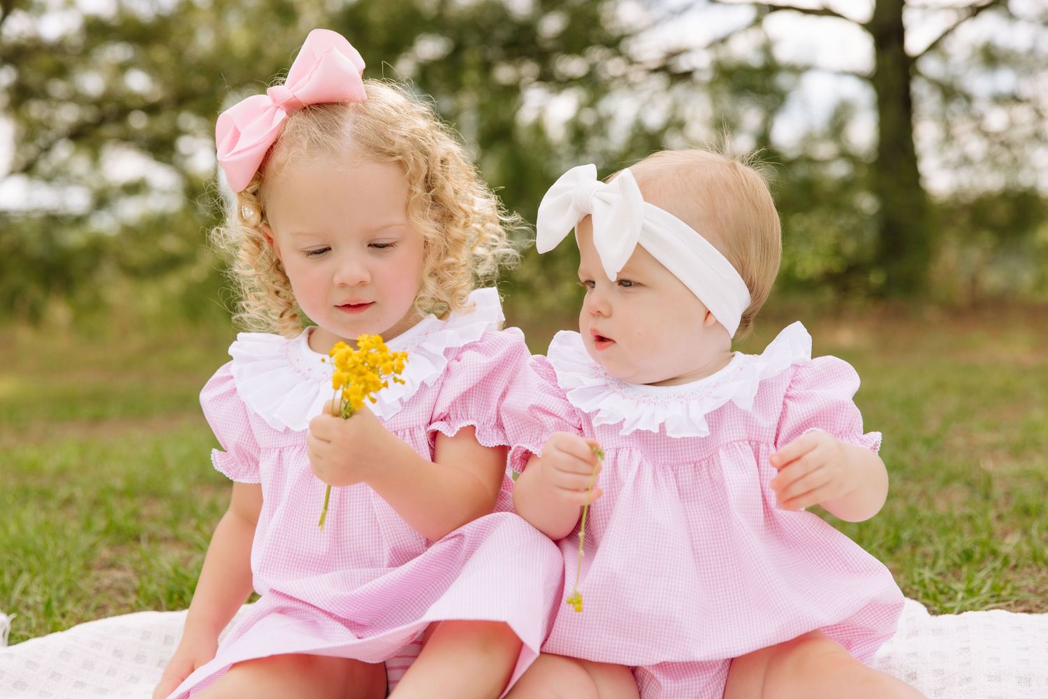 Two toddler girls in matching pink dresses sit together outdoors, one holding yellow flowers.