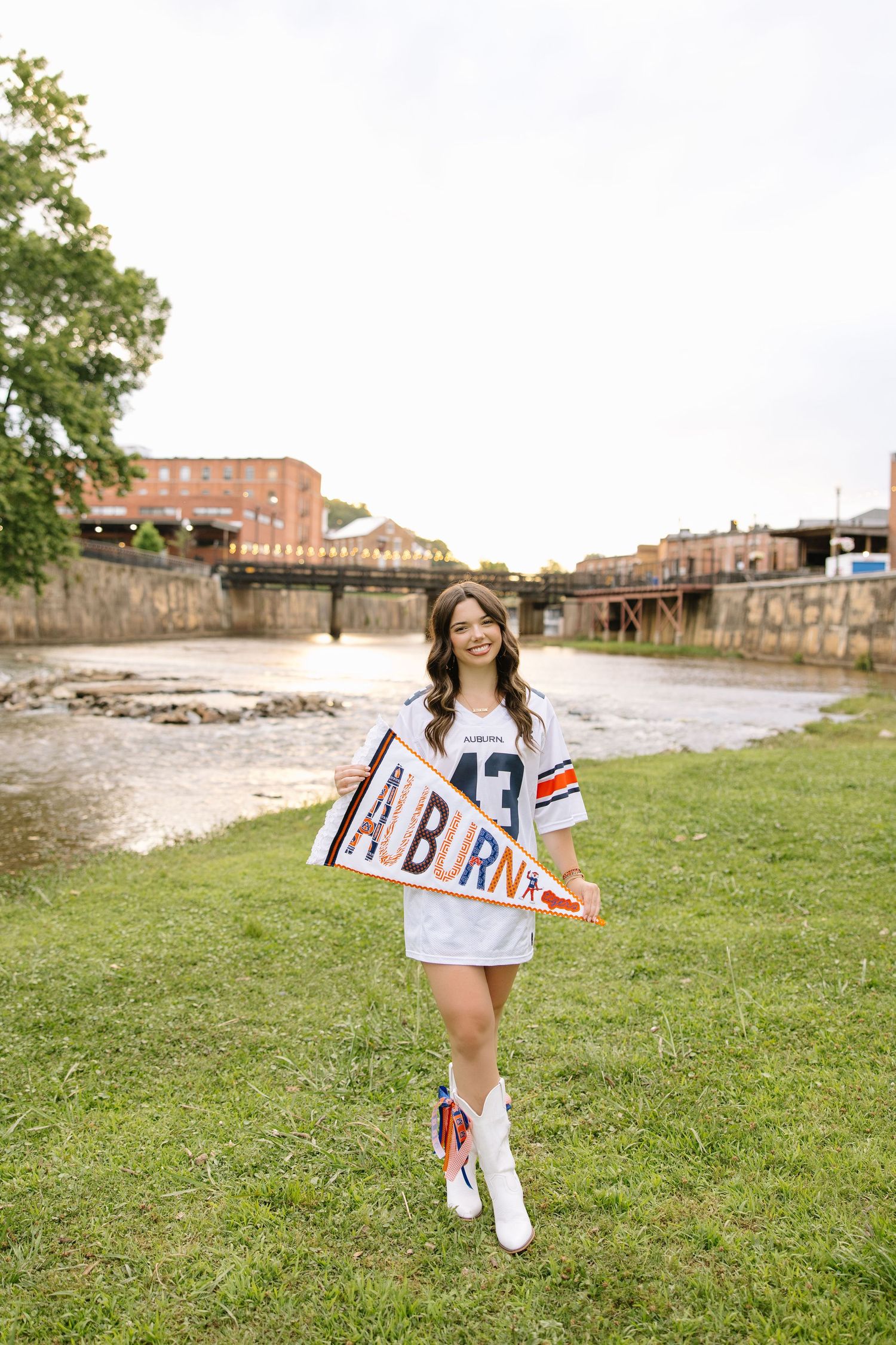 Young woman in Auburn jersey and white boots holds an Auburn pennant by a river on a cloudy day.