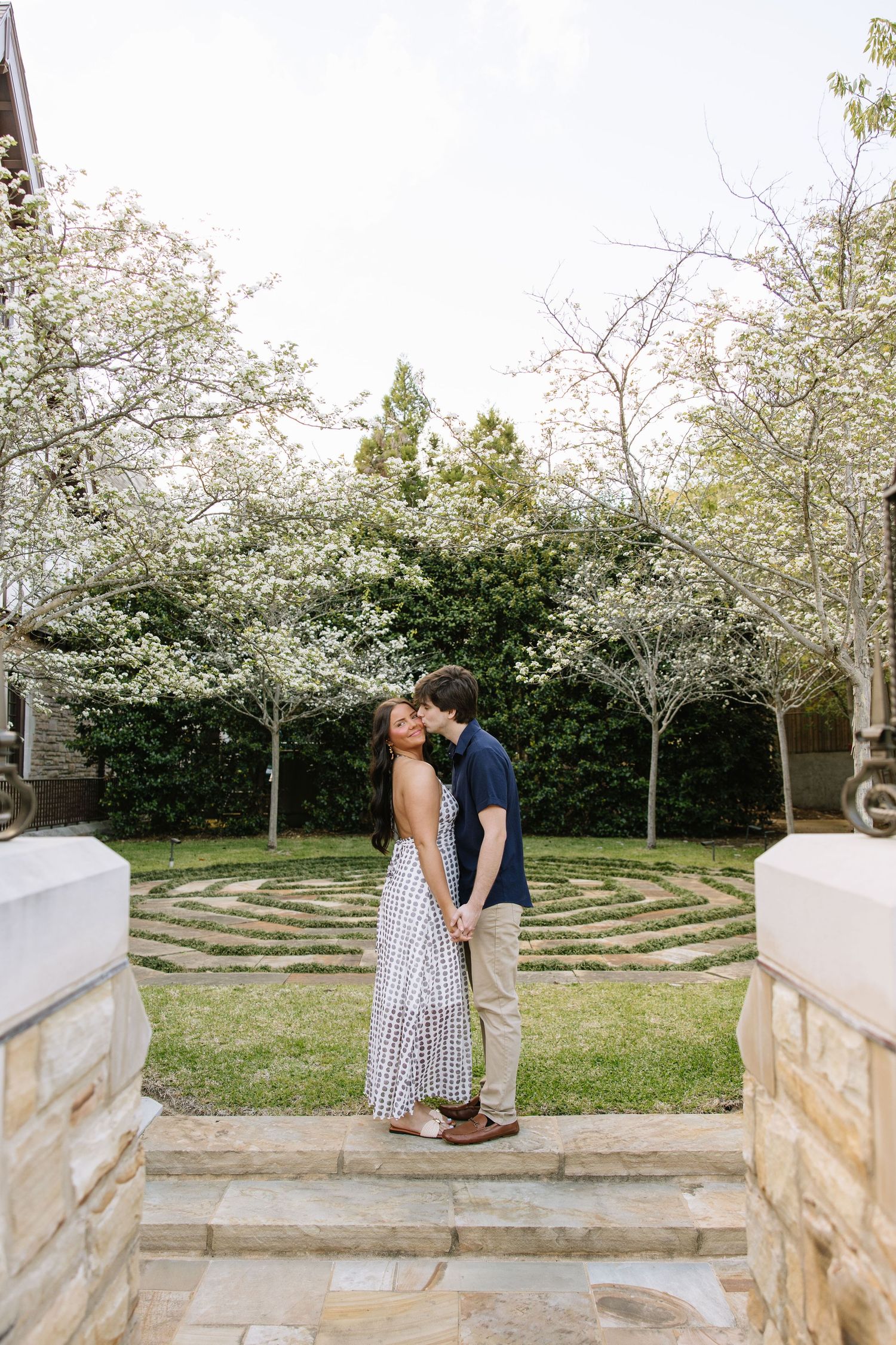 Couple holding hands and kissing in a garden with blooming white trees and a stone labyrinth backdrop.