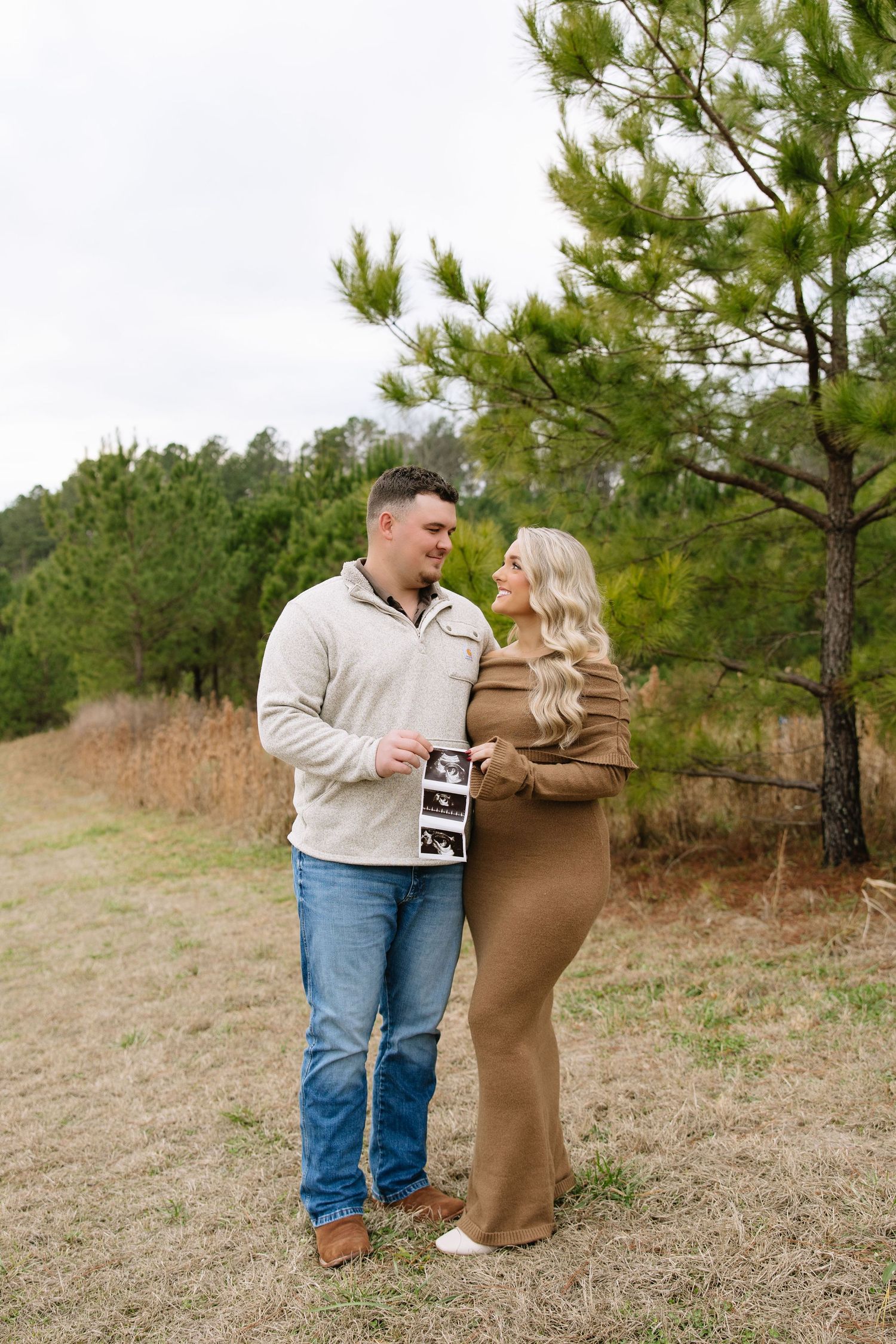 A couple stands outdoors holding ultrasound photos, smiling at each other near pine trees, announcing their pregnancy.
