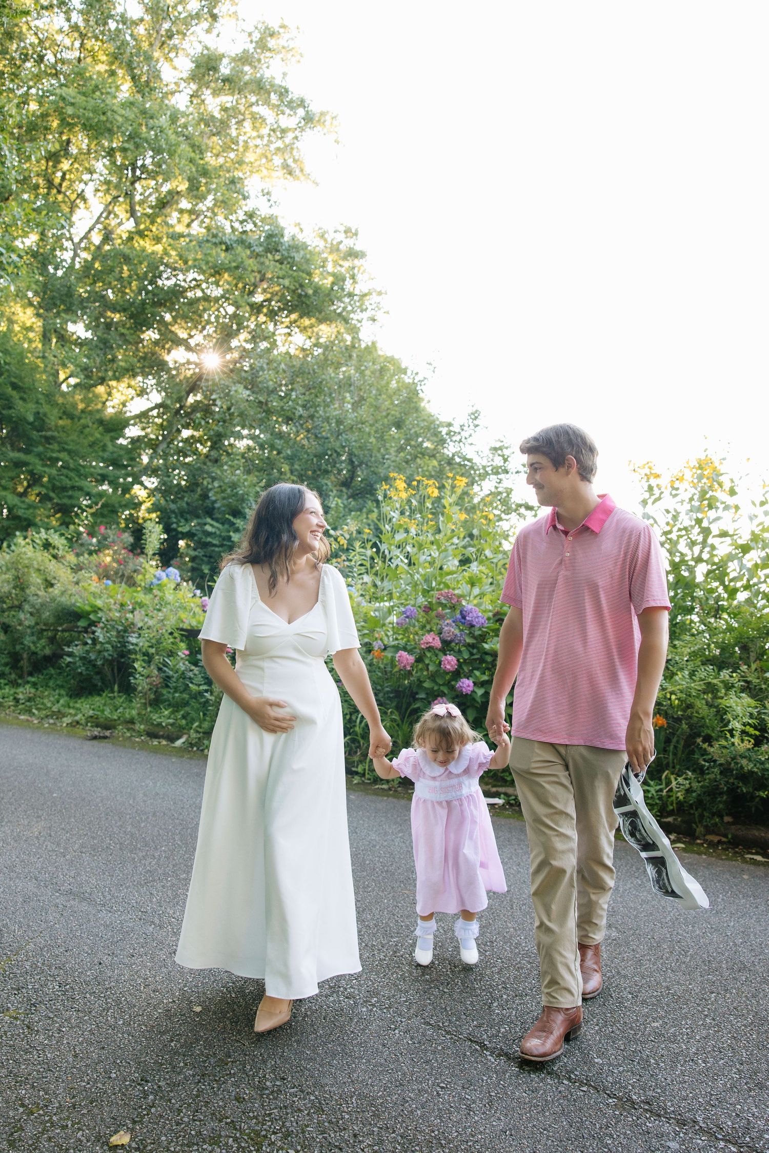 Pregnant woman in white dress walks with husband and toddler daughter in pink dress through a sunny garden path.