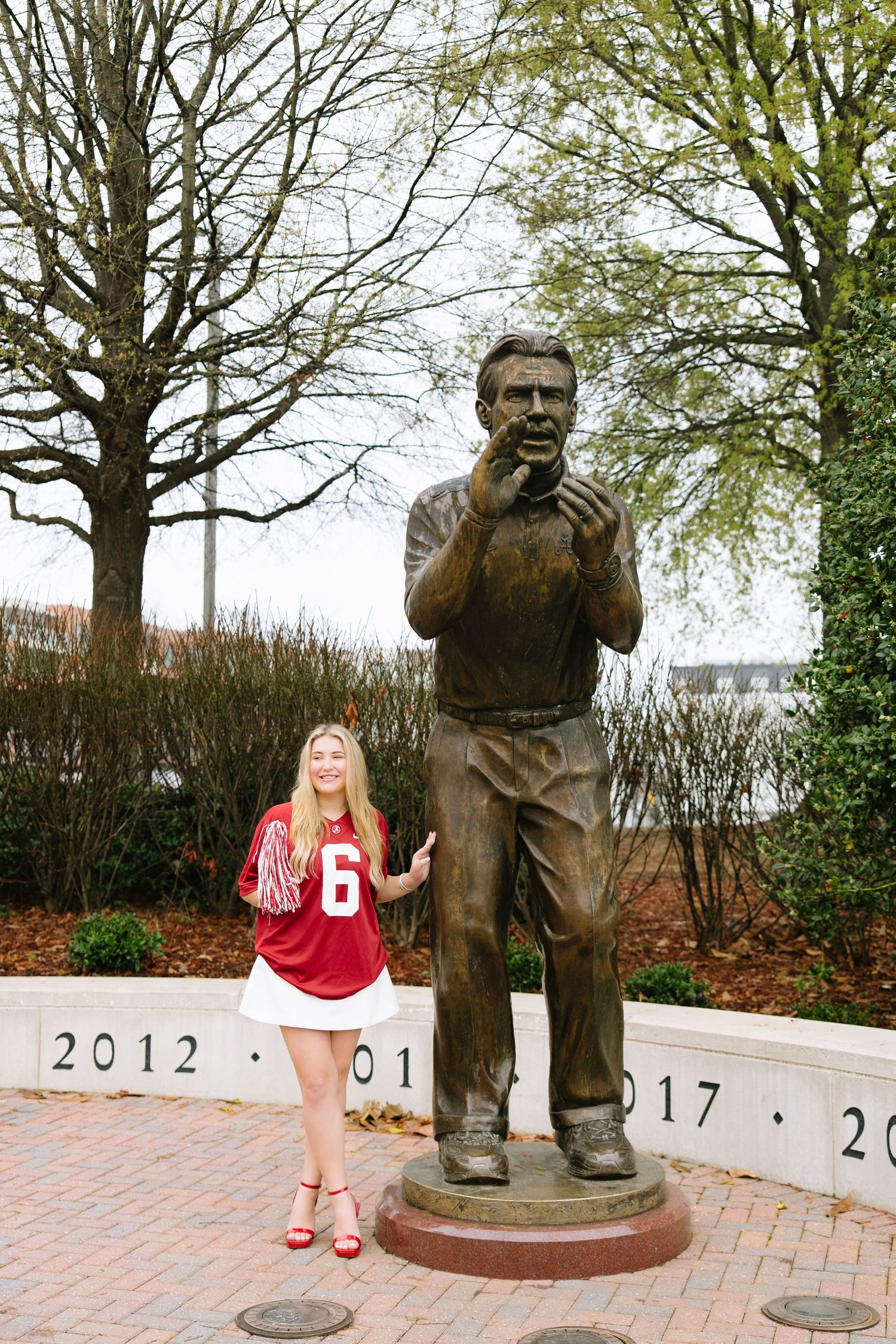 Young woman in Alabama Crimson Tide jersey poses beside a bronze coach statue on a brick plaza.