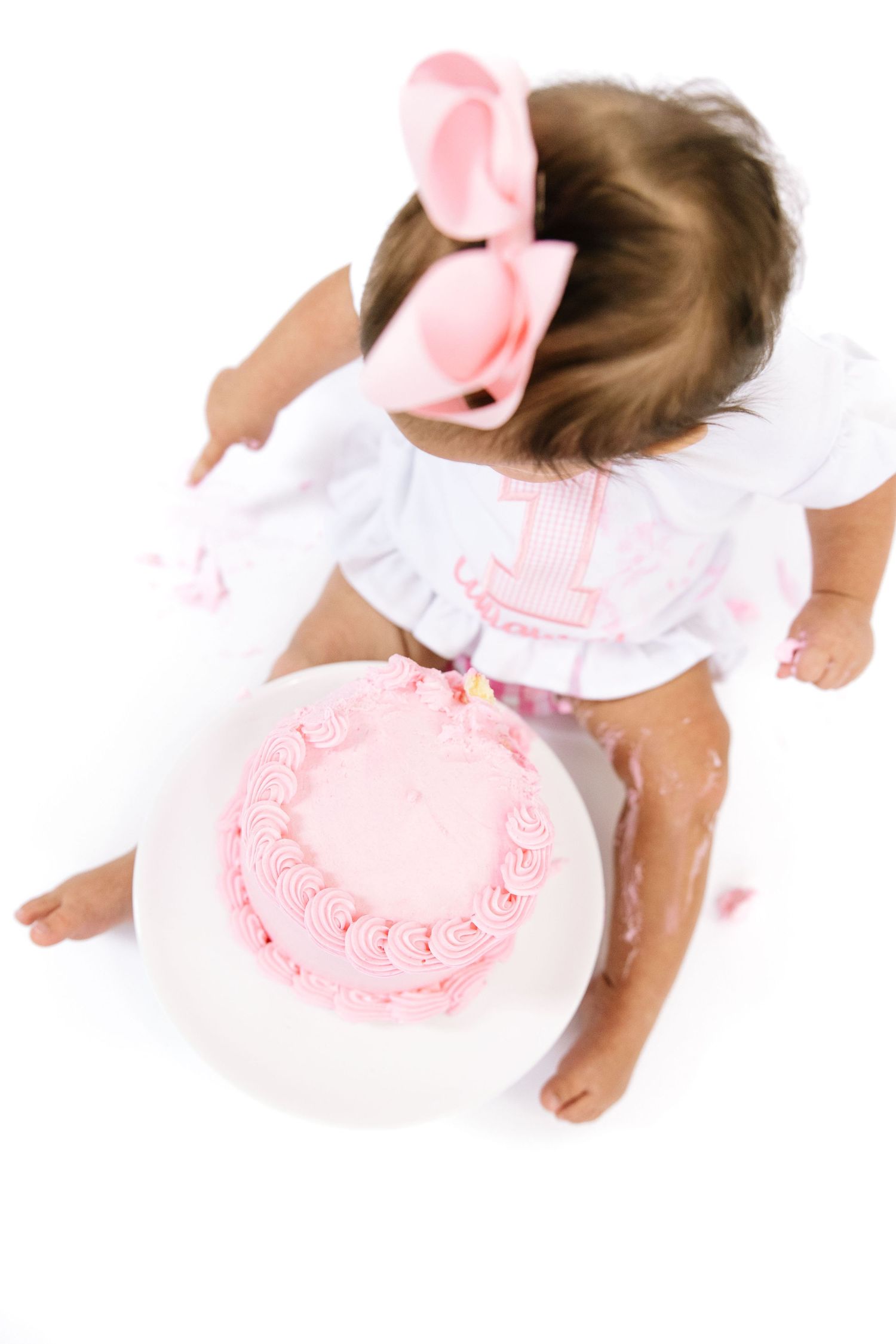 Overhead view of baby girl in white dress with pink bow reaching for her first birthday smash cake.