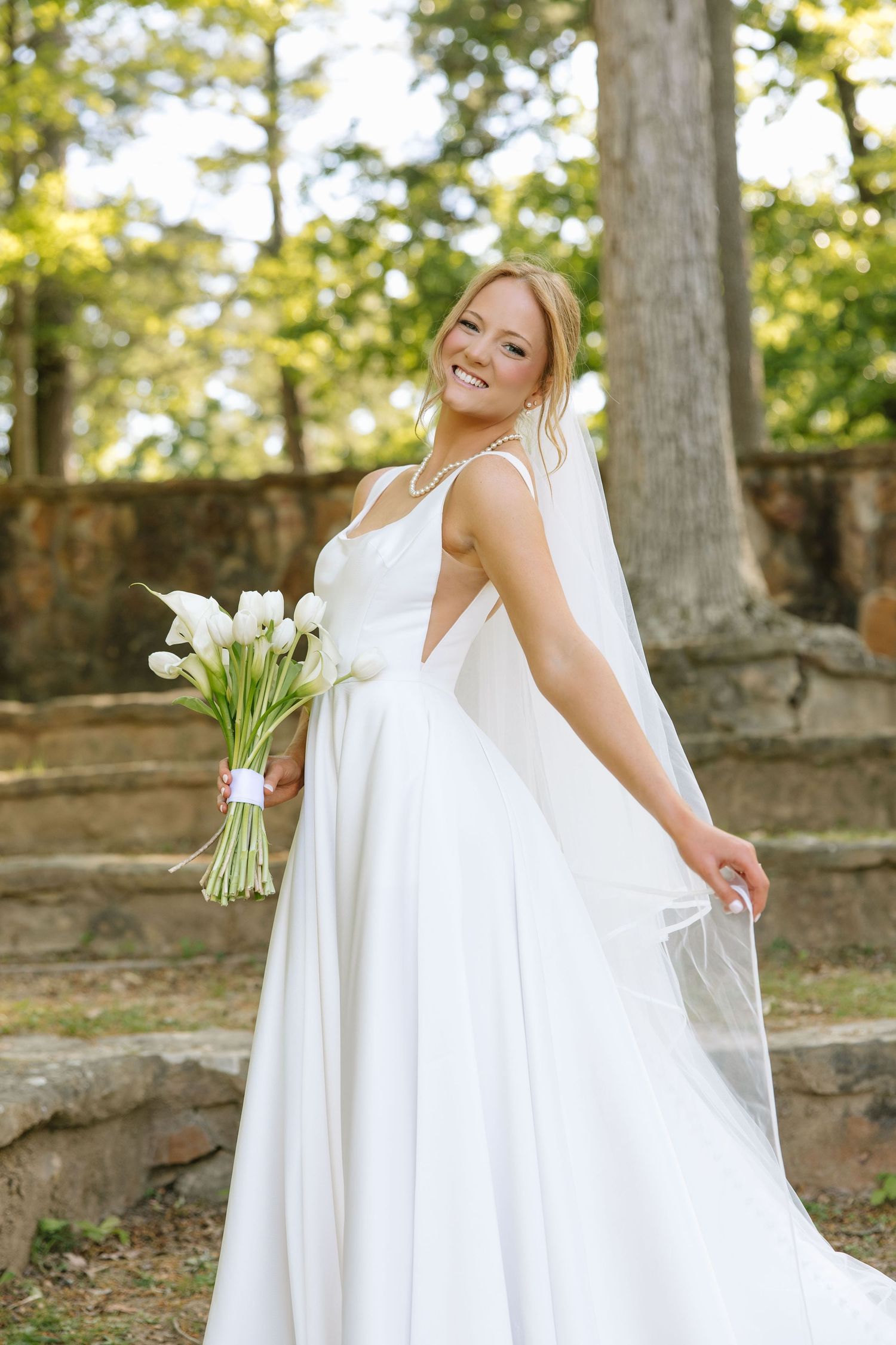 Smiling bride in white gown holds tulip bouquet, veil flowing, posing outdoors among trees and stone steps.