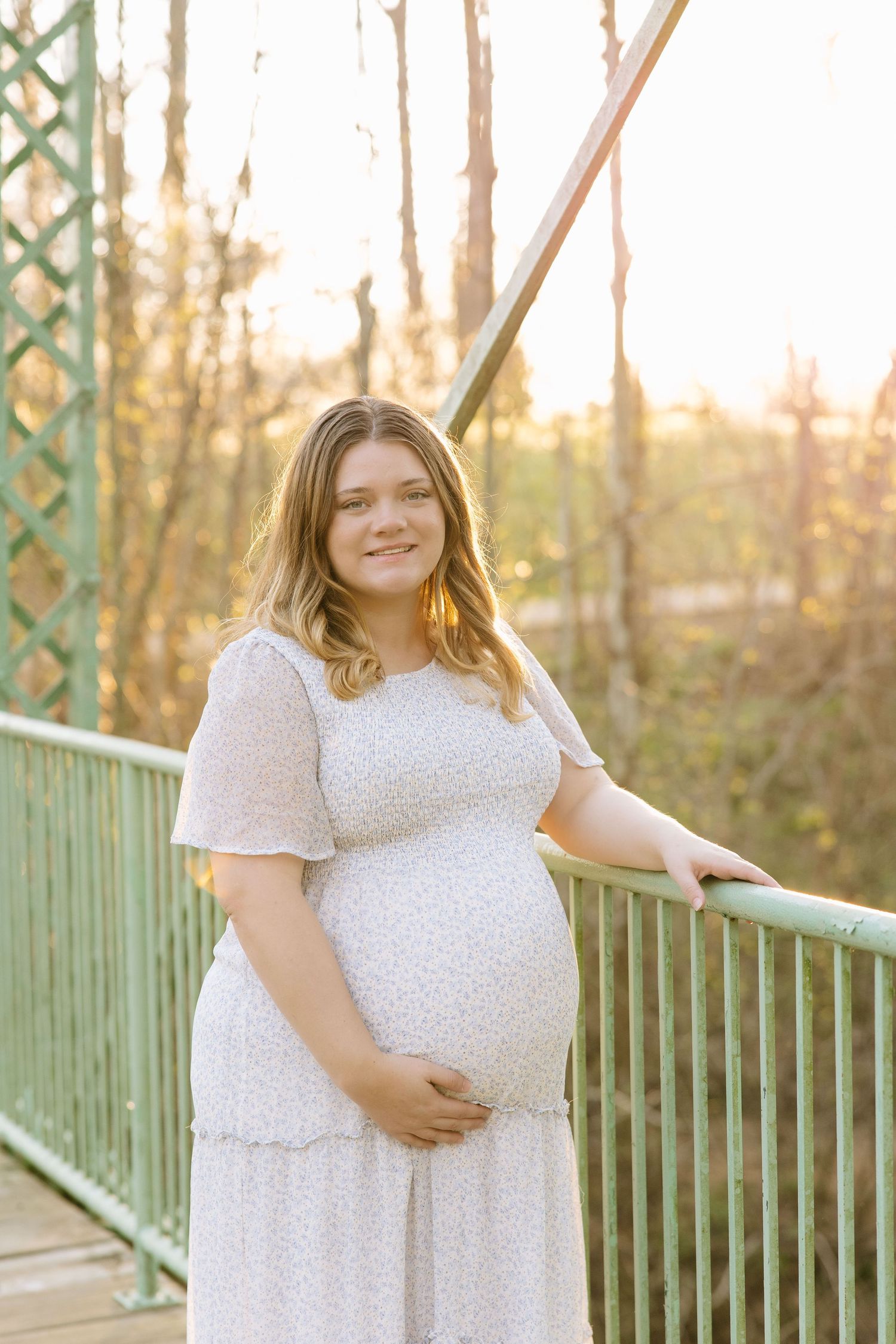 Pregnant woman in white floral dress smiling on a green bridge surrounded by trees in golden hour light.
