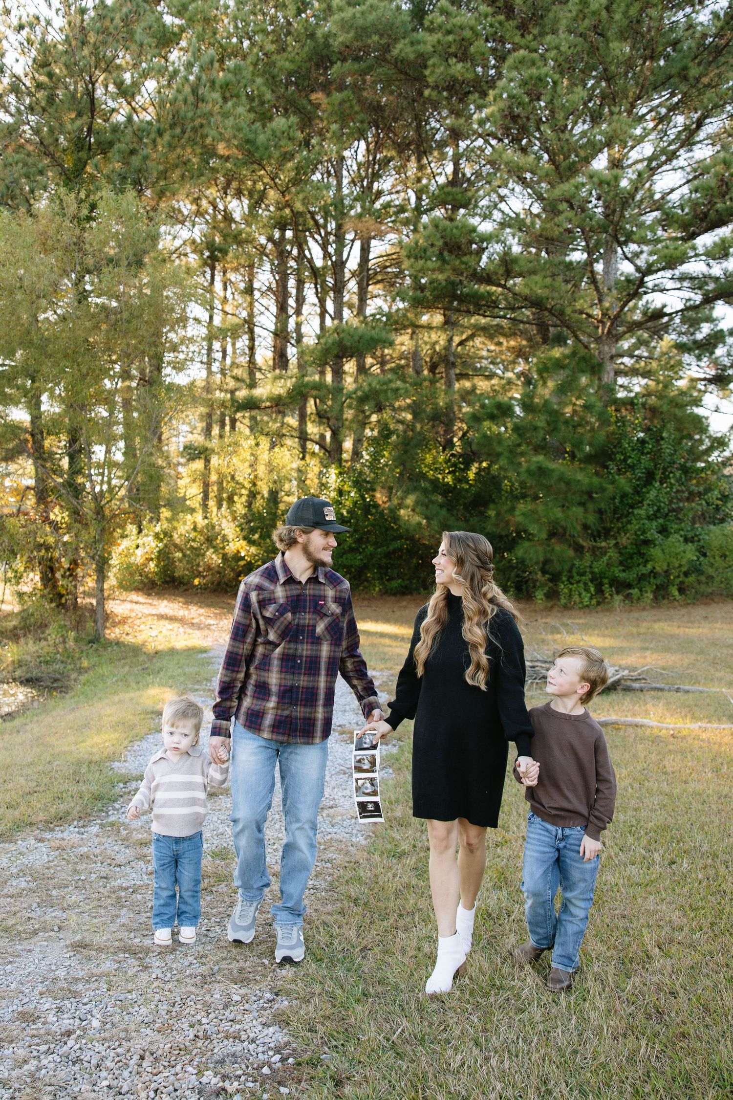 Family of four walking outdoors, holding ultrasound photos, announcing pregnancy among tall pine trees in autumn.