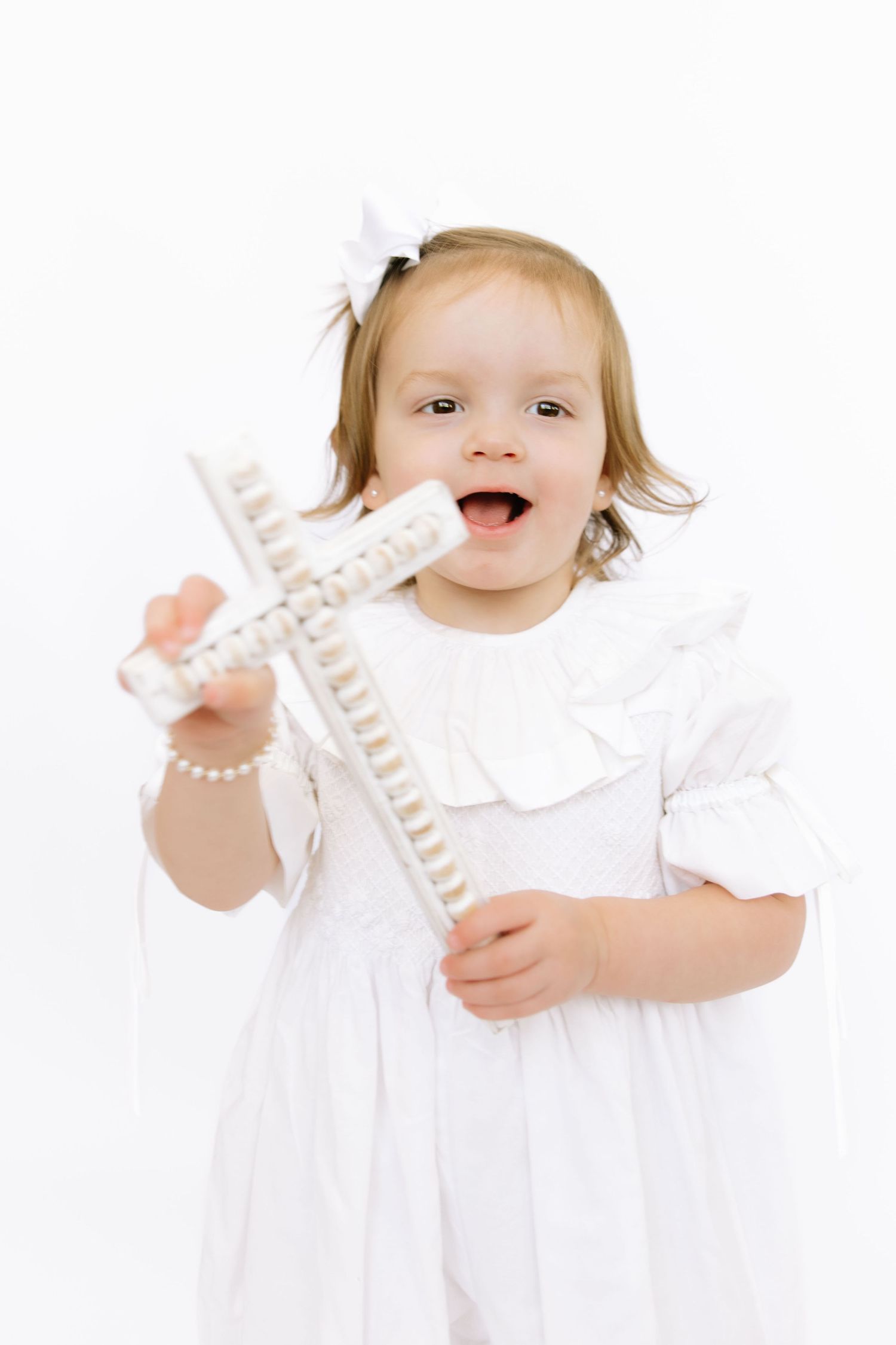 Happy toddler girl in white dress holding a decorative cross, smiling joyfully against white background.