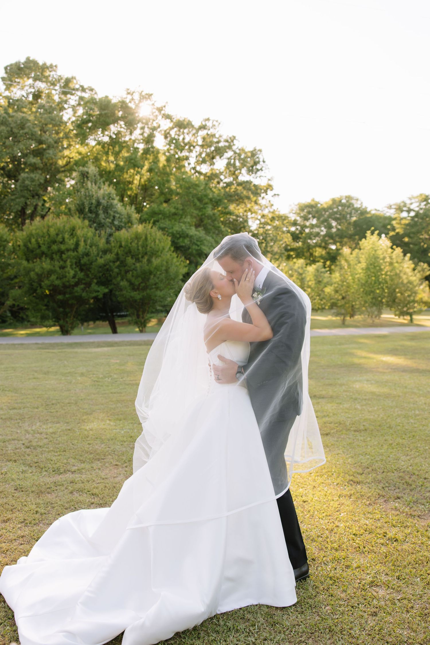 Bride and groom kissing under a flowing white veil at golden hour in a lush green park setting.