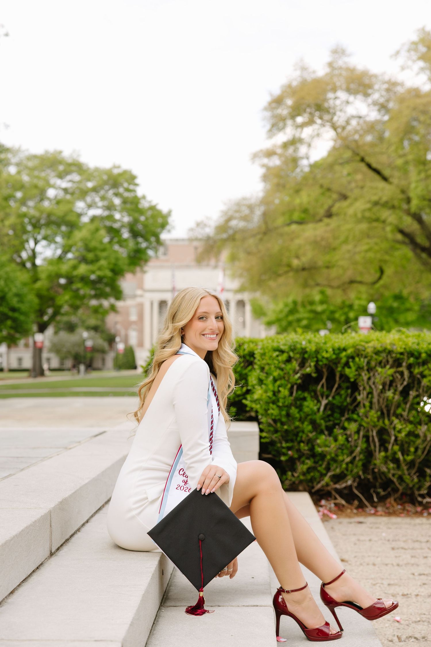 Smiling blonde woman in white dress holding graduation cap on campus steps wearing red heels.