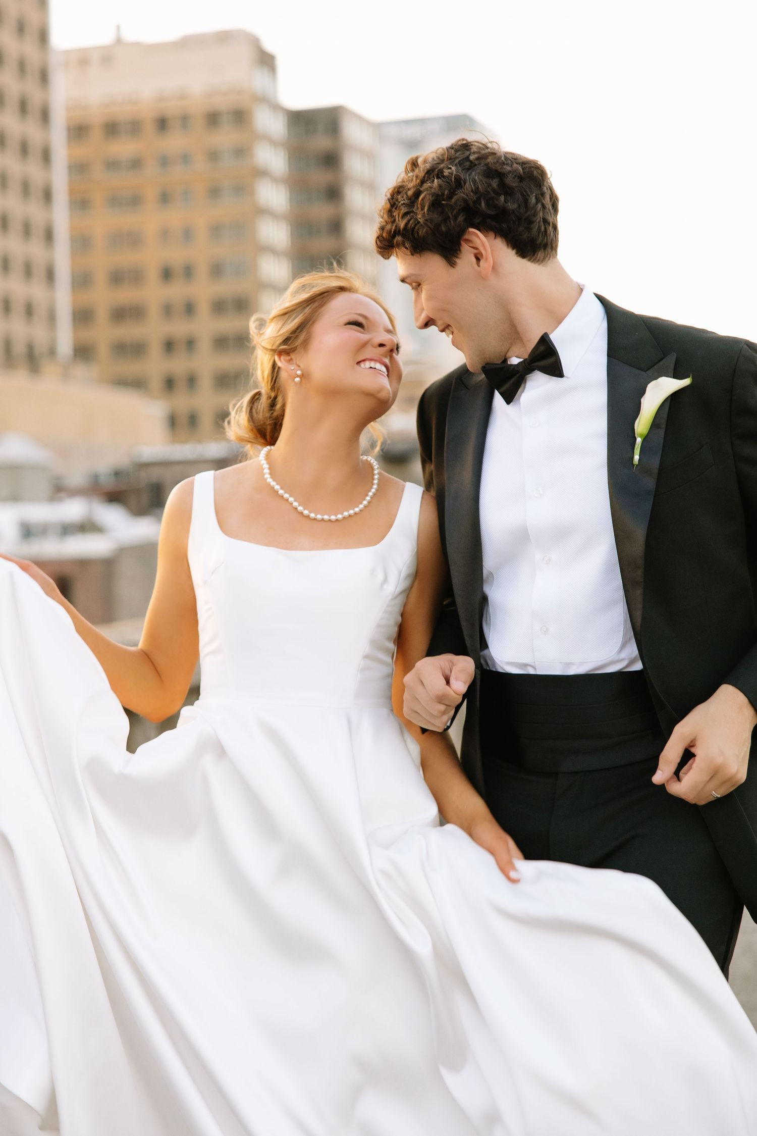 Joyful bride and groom walk together on a rooftop, bride lifting her white gown with city views.