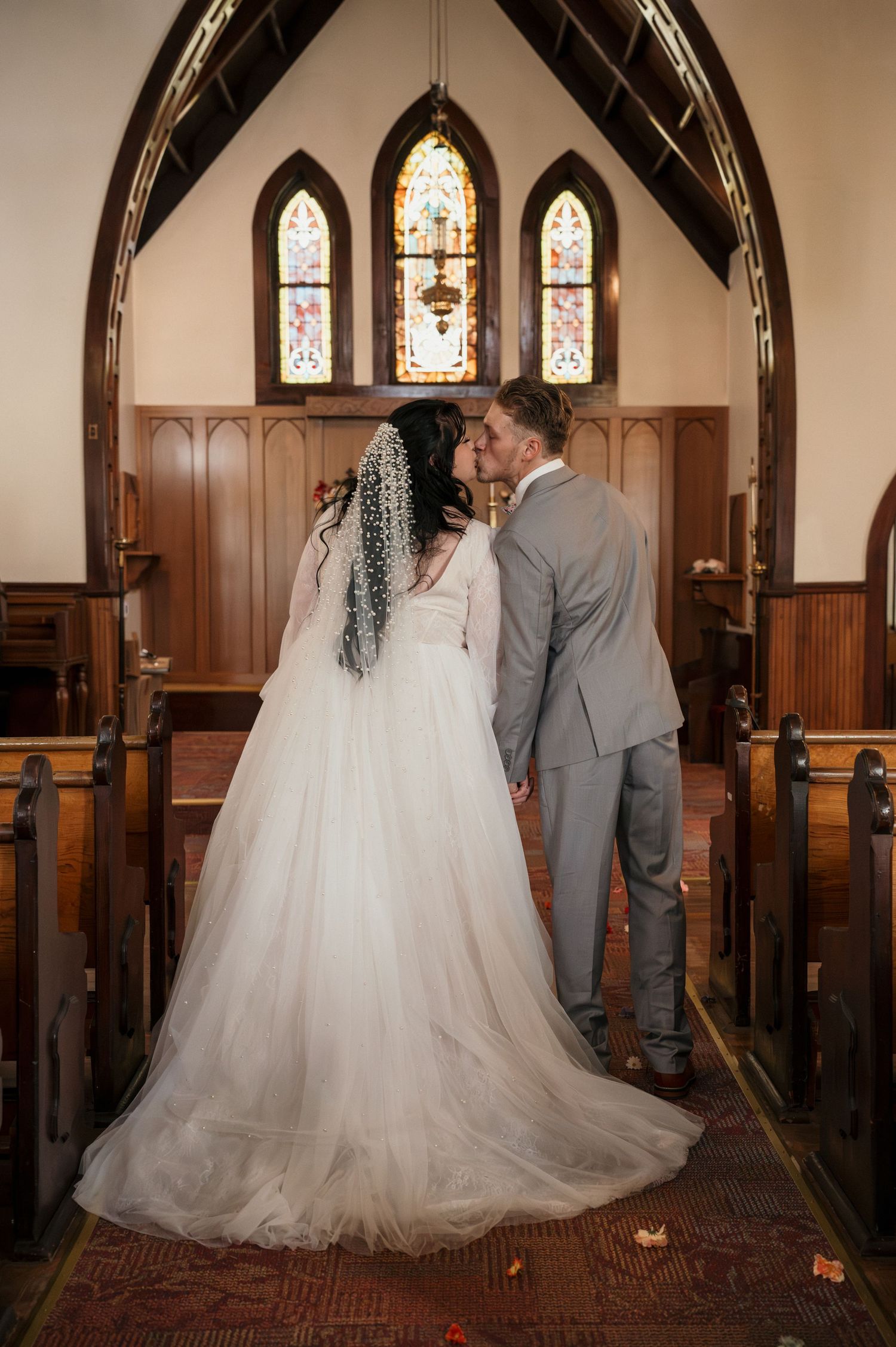A bride and a groom sharing a kiss in the aisle of a dark beautiful church with stained glass windows in Payette, Idaho.
