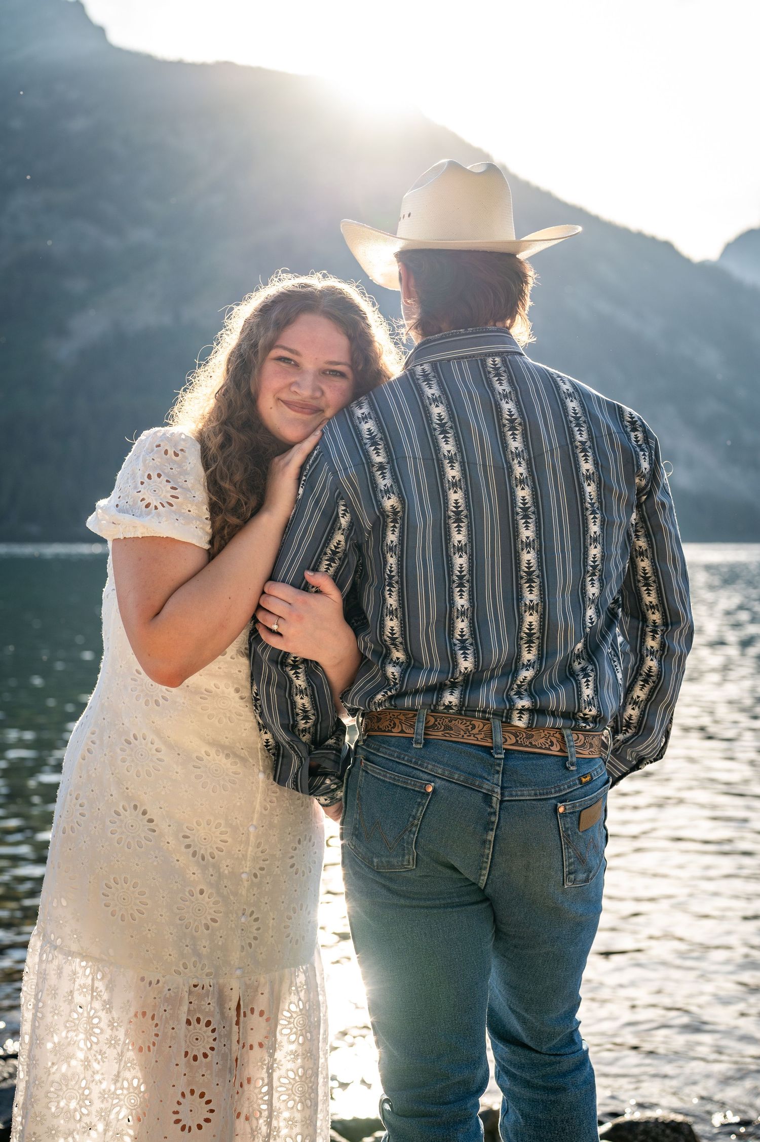 A girl looks at the camera over the shoulder of her cowboy fiancé at Jenny Lake in Grand Teton National Park.