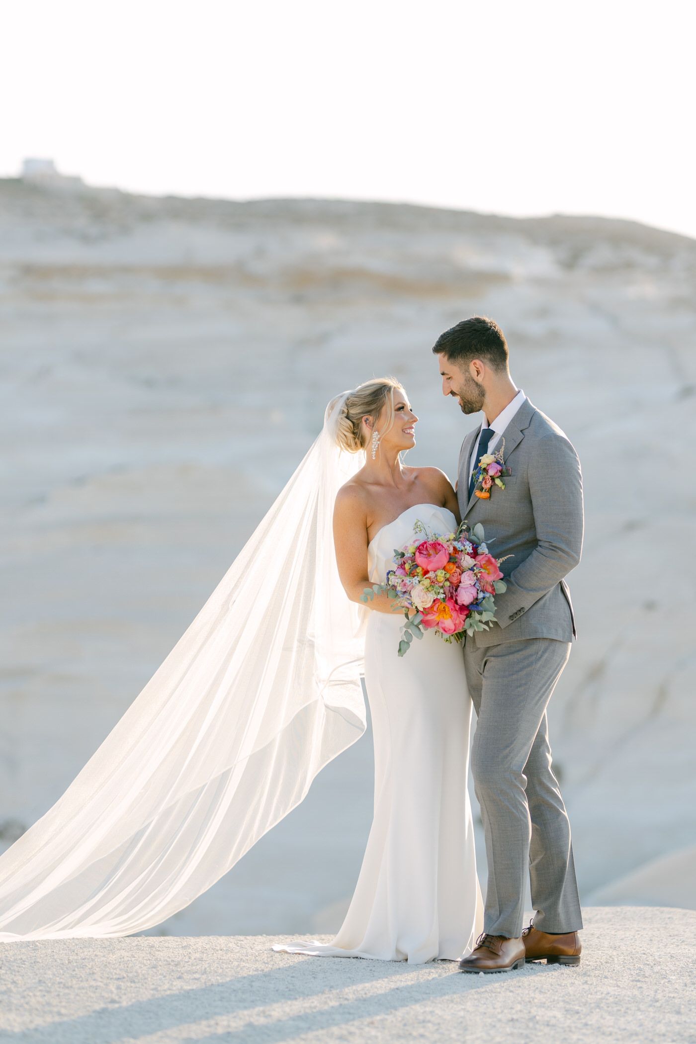 A bride in white gown with long veil and groom in grey suit share a moment while holding pink bouquet in Milos Island