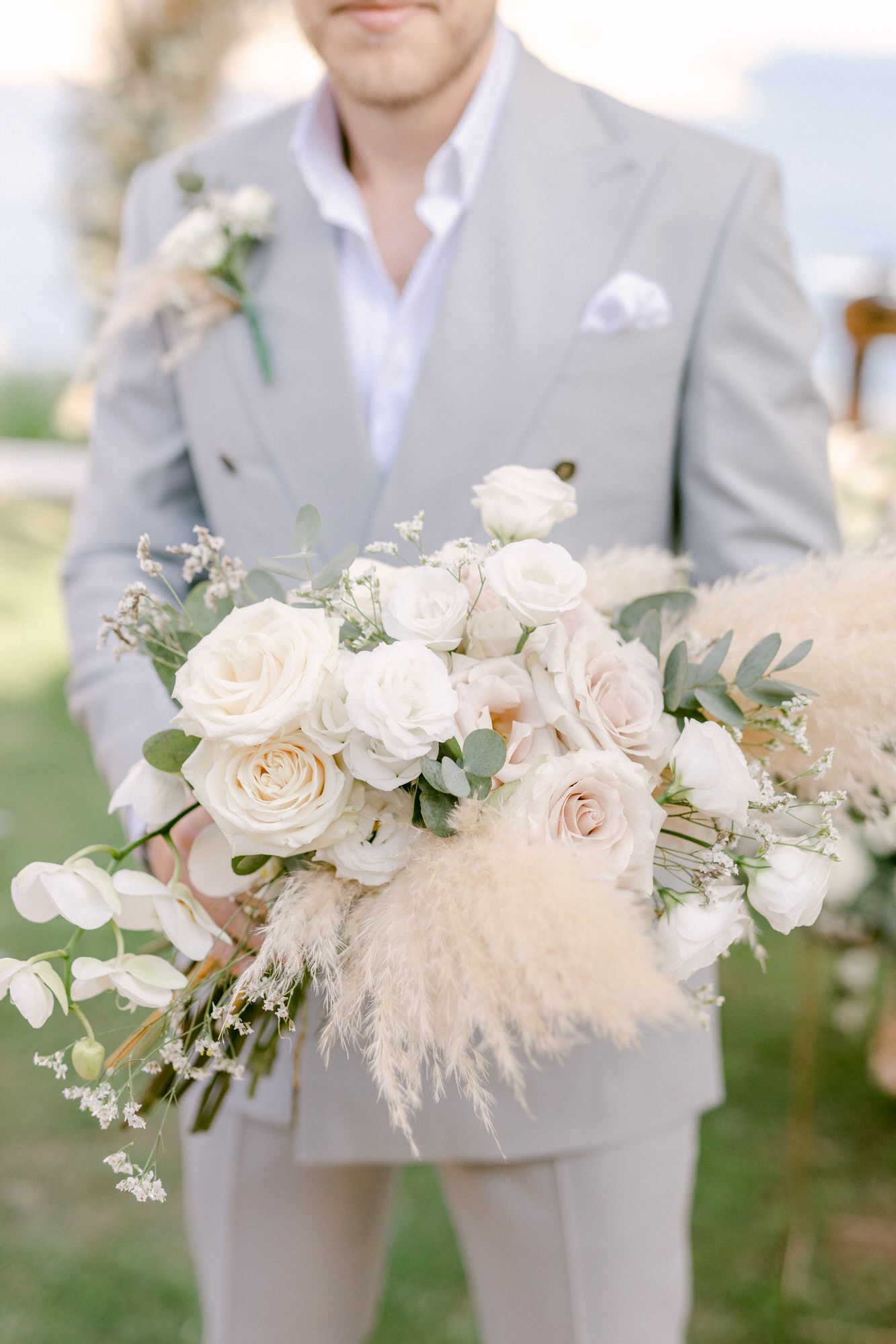Elegant white and blush wedding bouquet with roses and pampas grass held against grey suit.