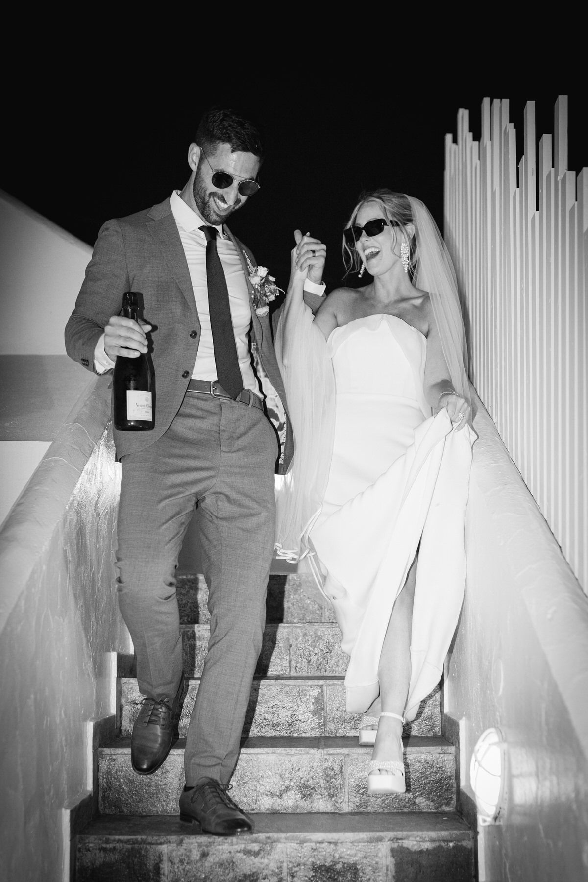 A black and white photo of a newlywed couple having fun on steps, with the groom holding a bottle and both striking poses.