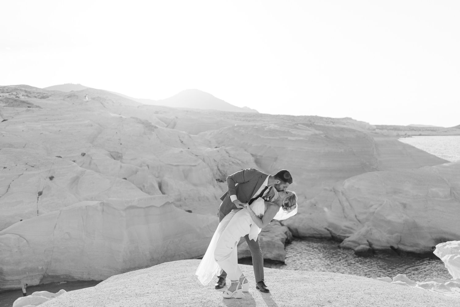 A couple in wedding attire embraces against a dramatic desert rock formation backdrop in black and white.