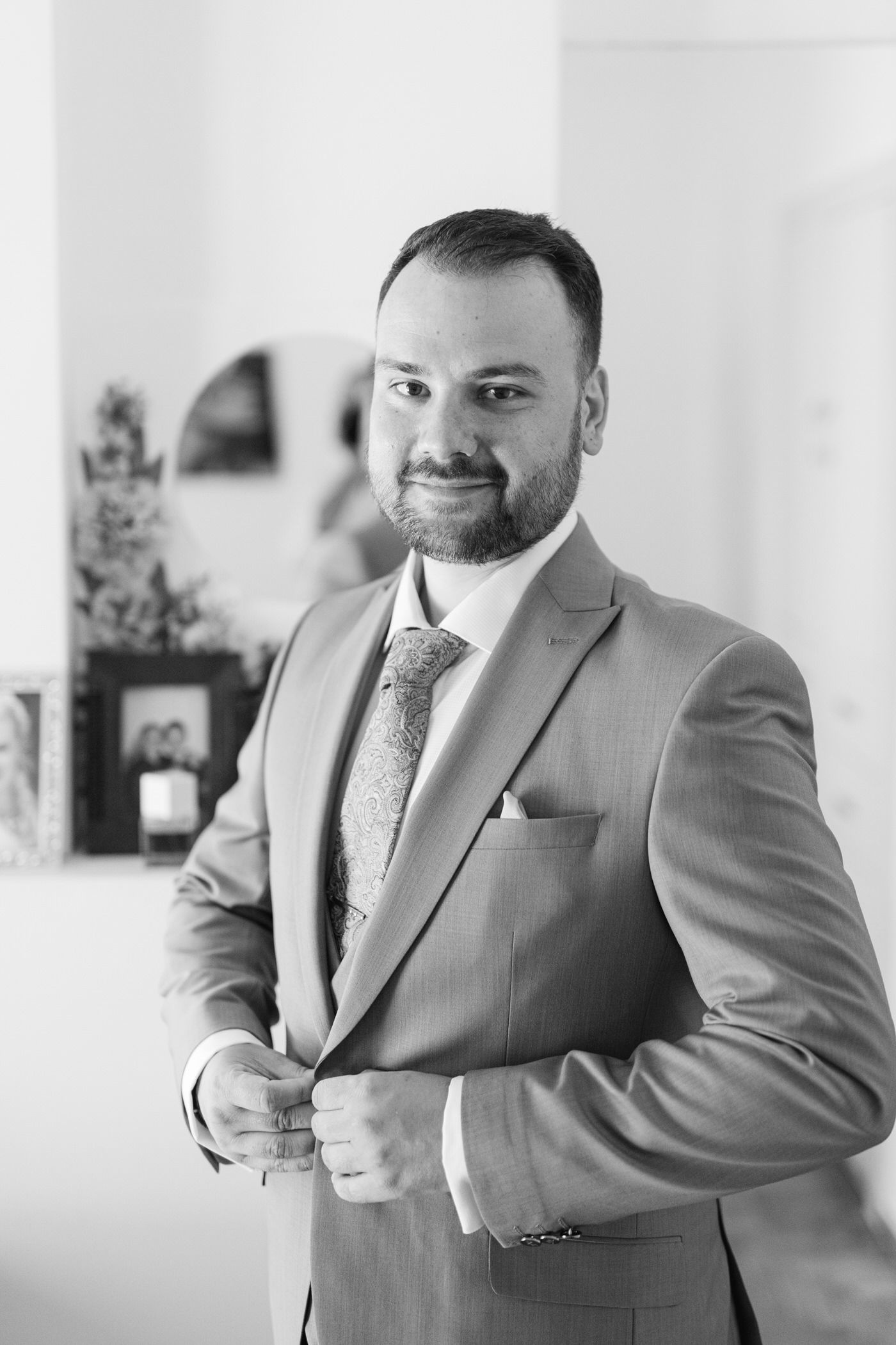 A professional portrait of a bearded man in a gray suit and patterned tie, smiling at the camera in black and white.