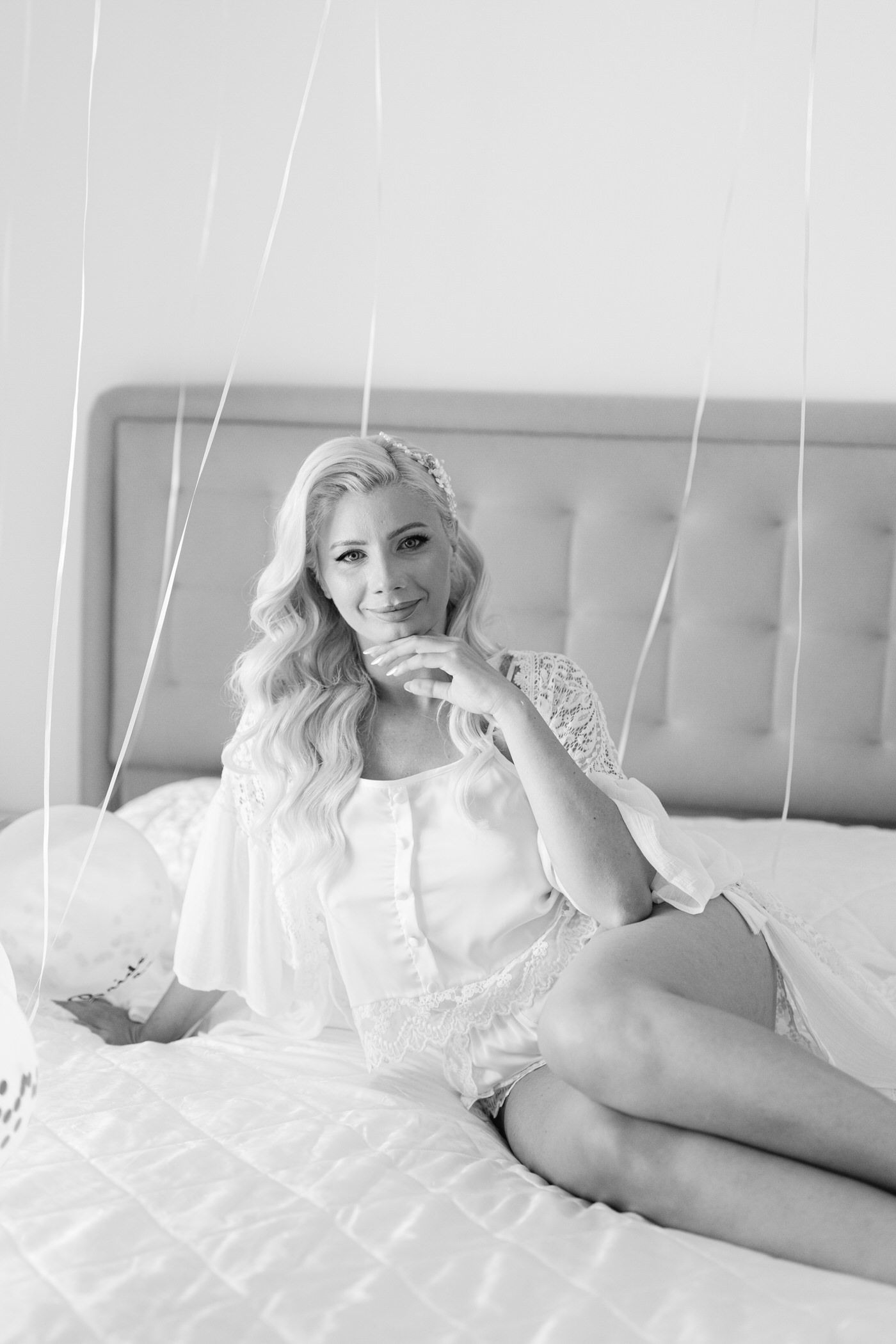 Black and white boudoir photo showing woman in white lace loungewear reclining on tufted headboard bed.