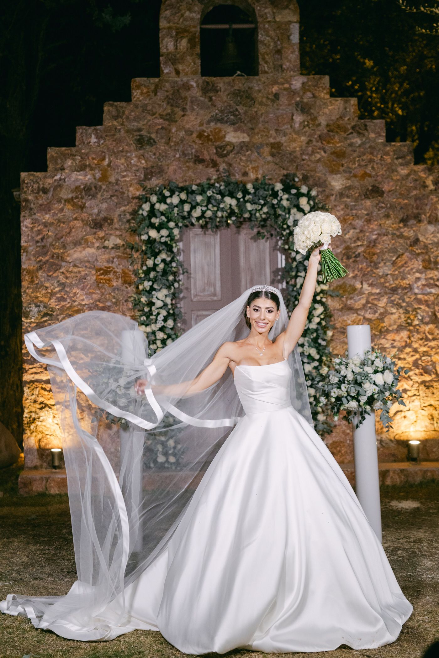 A flowing white wedding gown is captured at night in front of a stone archway decorated with floral garlands and lights.