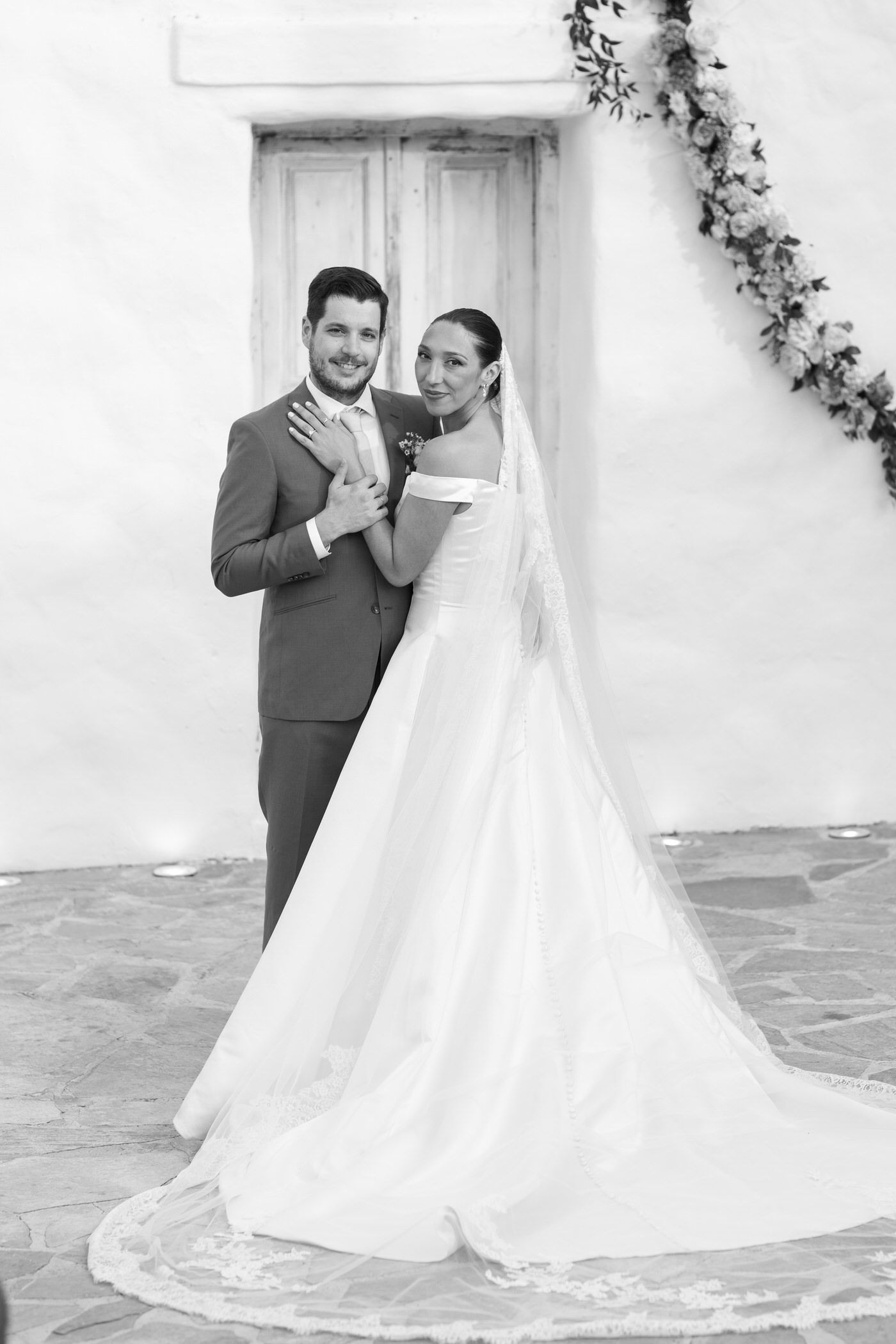 A black and white wedding portrait showing a couple in formal attire posing in front of a white wall with climbing vines.