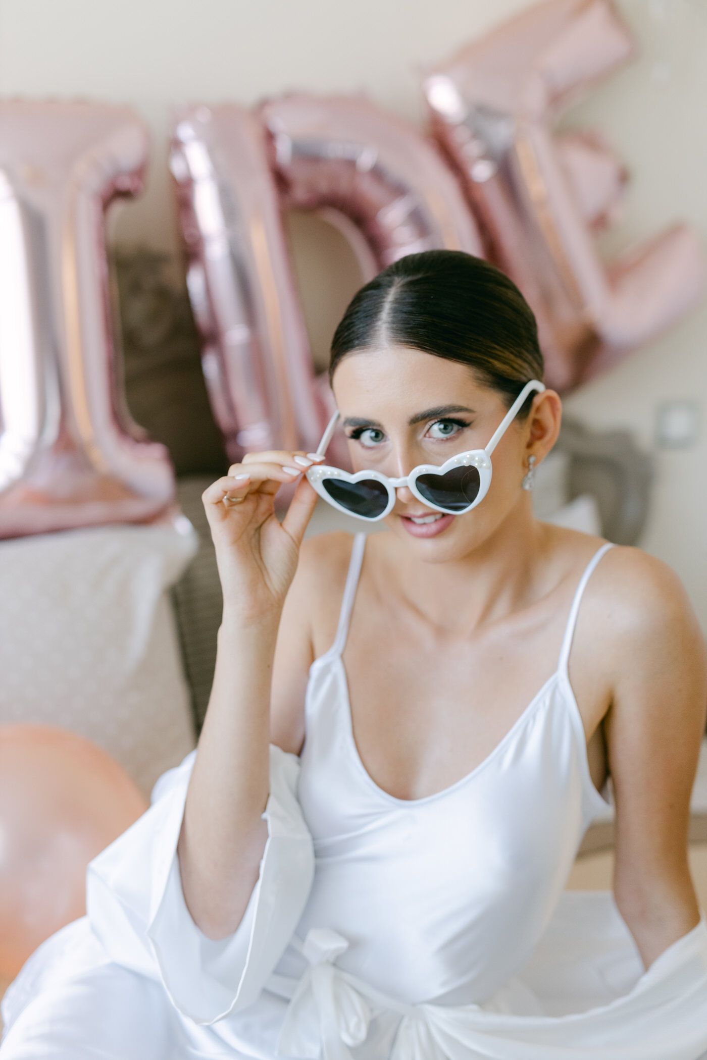 Woman wearing white dress playfully poses with heart-shaped sunglasses prop against rose gold balloon decor.