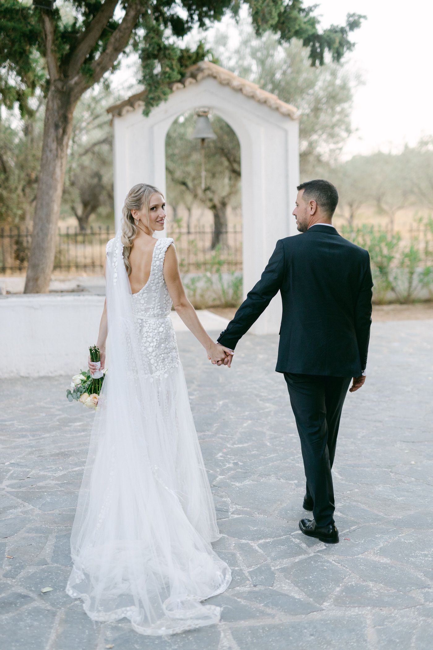 A couple in wedding attire holding hands near a white arched entrance with lush greenery in the background.