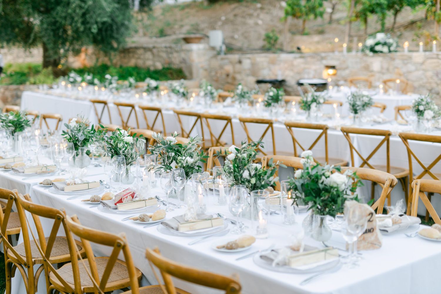Elegant outdoor wedding reception table with wooden crossback chairs and delicate floral centerpieces on white linens.