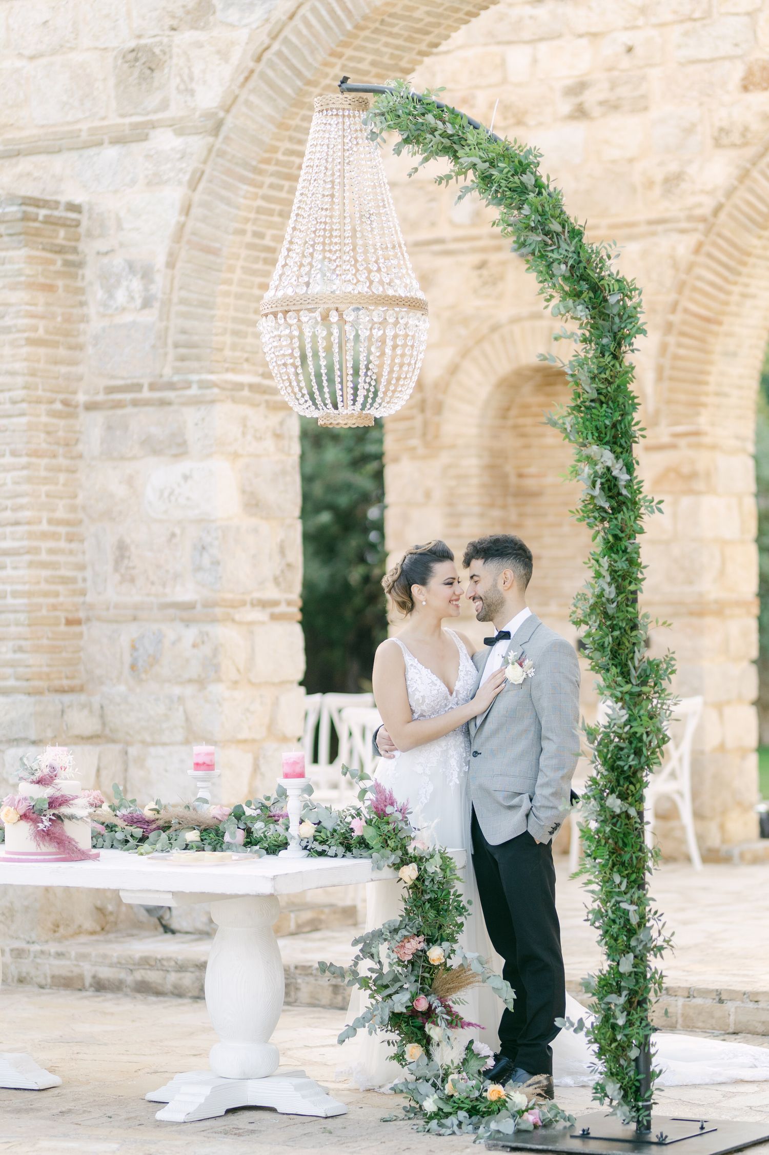 Romantic couple stands beneath crystal chandelier and greenery arch at stone venue.