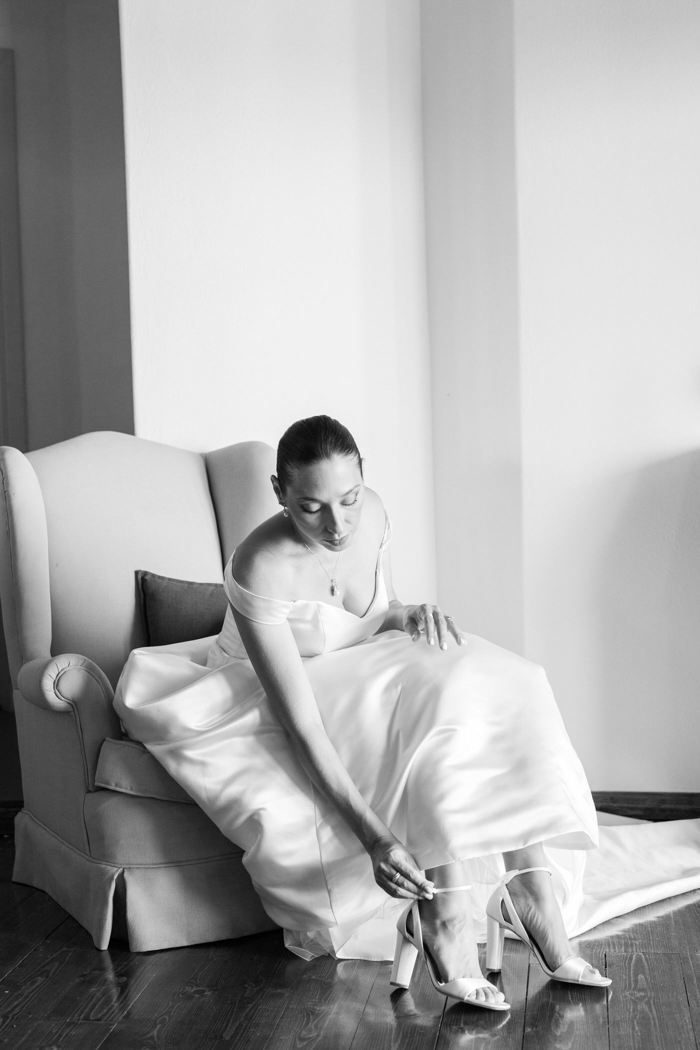 A woman in a white wedding gown sits elegantly in an armchair while adjusting her shoe in a black and white photograph.