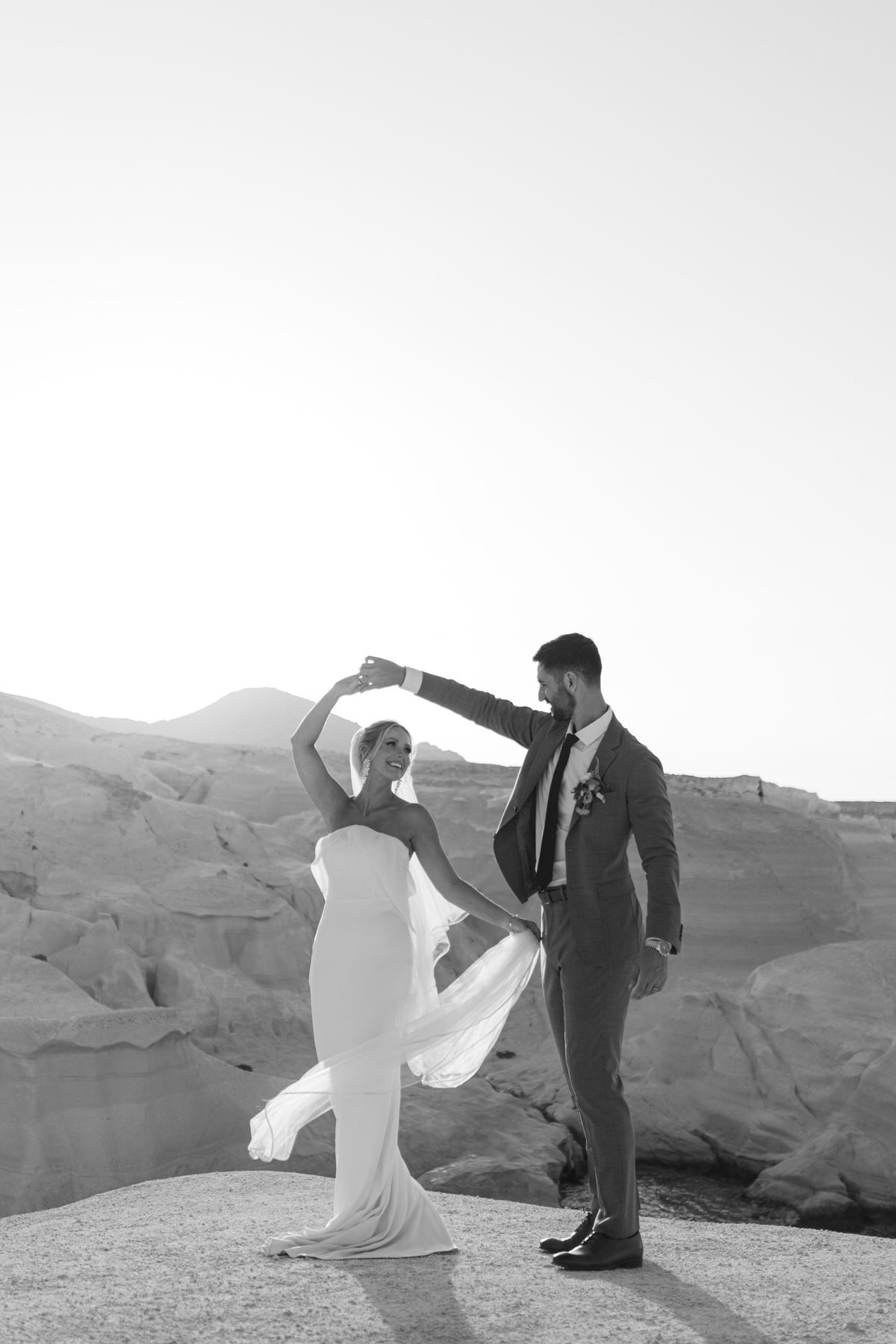 A couple dances atop a rocky mountain overlook in formal wedding attire, creating a dramatic black and white silhouette.