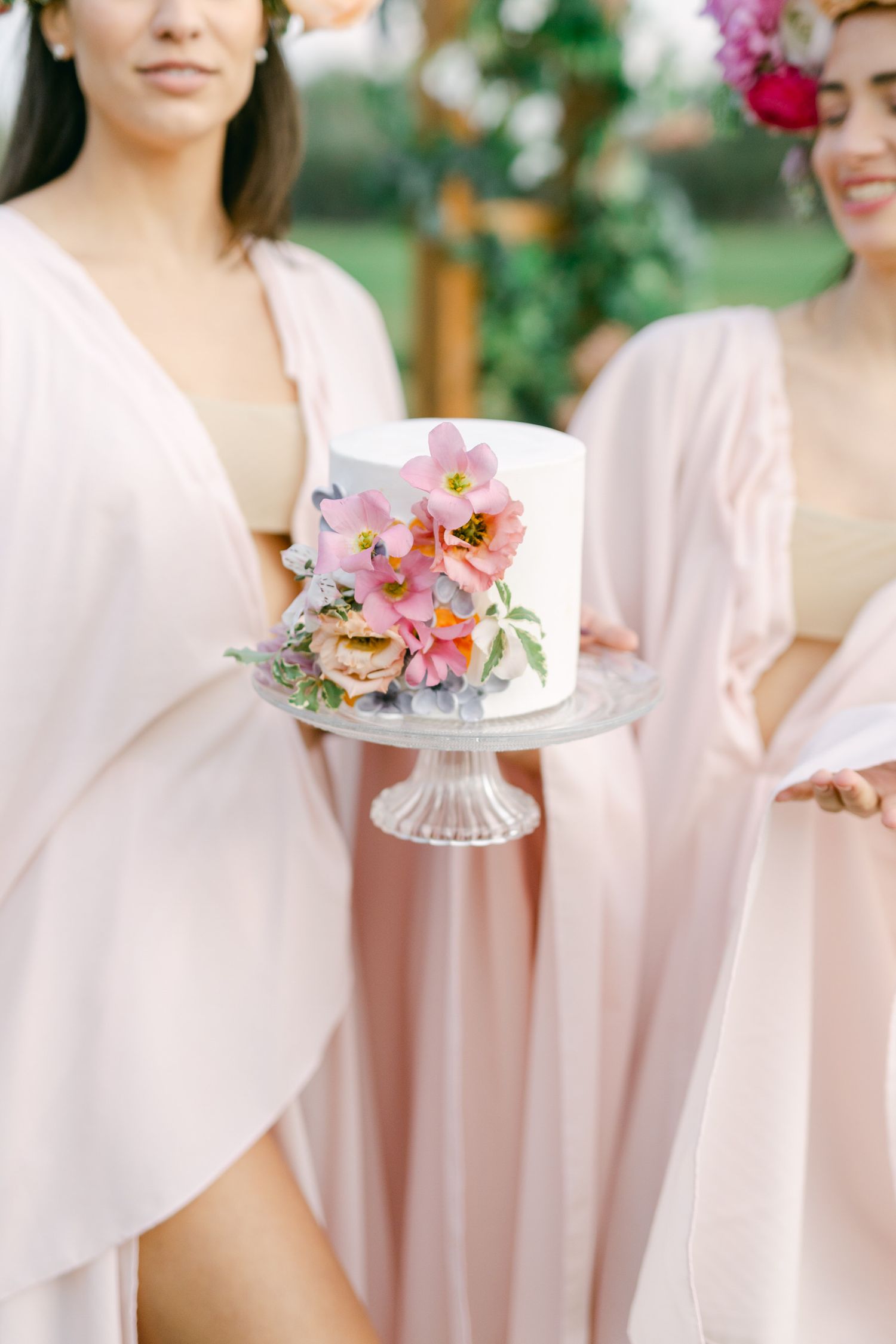 Small white wedding cake decorated with pink orchids and roses sits on crystal cake stand surrounded by blush pink dresses.
