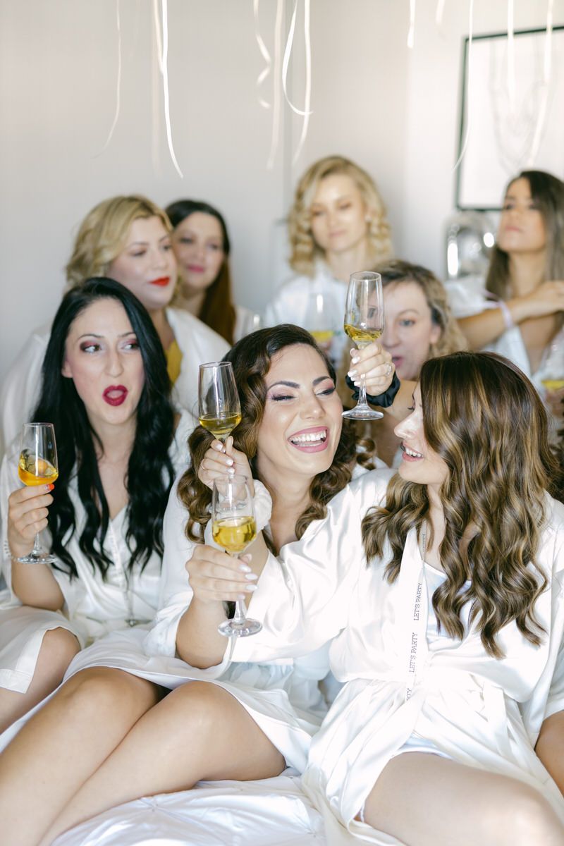 A group of friends in white robes holding glasses of champagne while celebrating together on a bed.