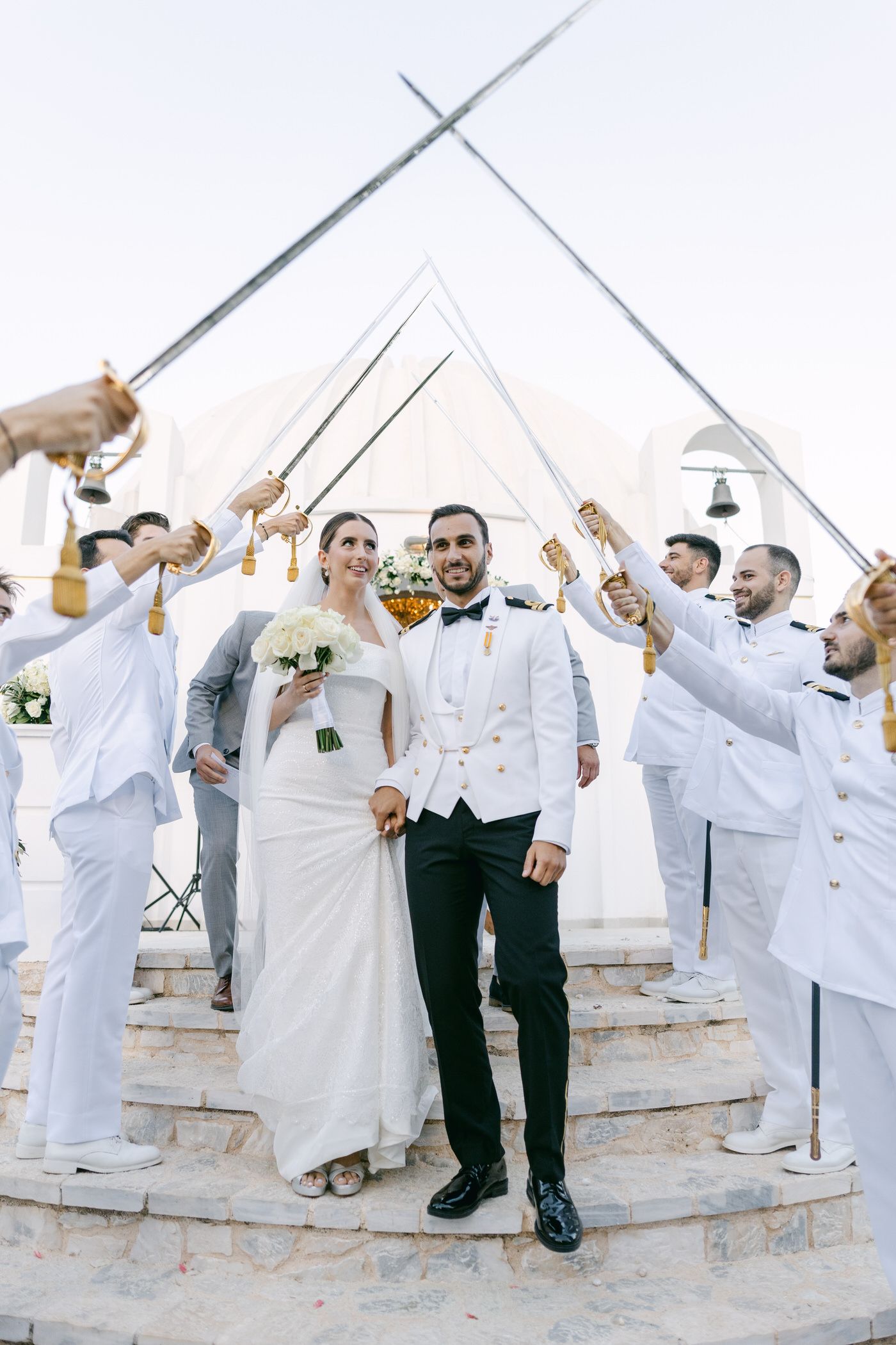 A newlywed couple descending white stone steps while guests in white outfits raise champagne glasses in celebration.