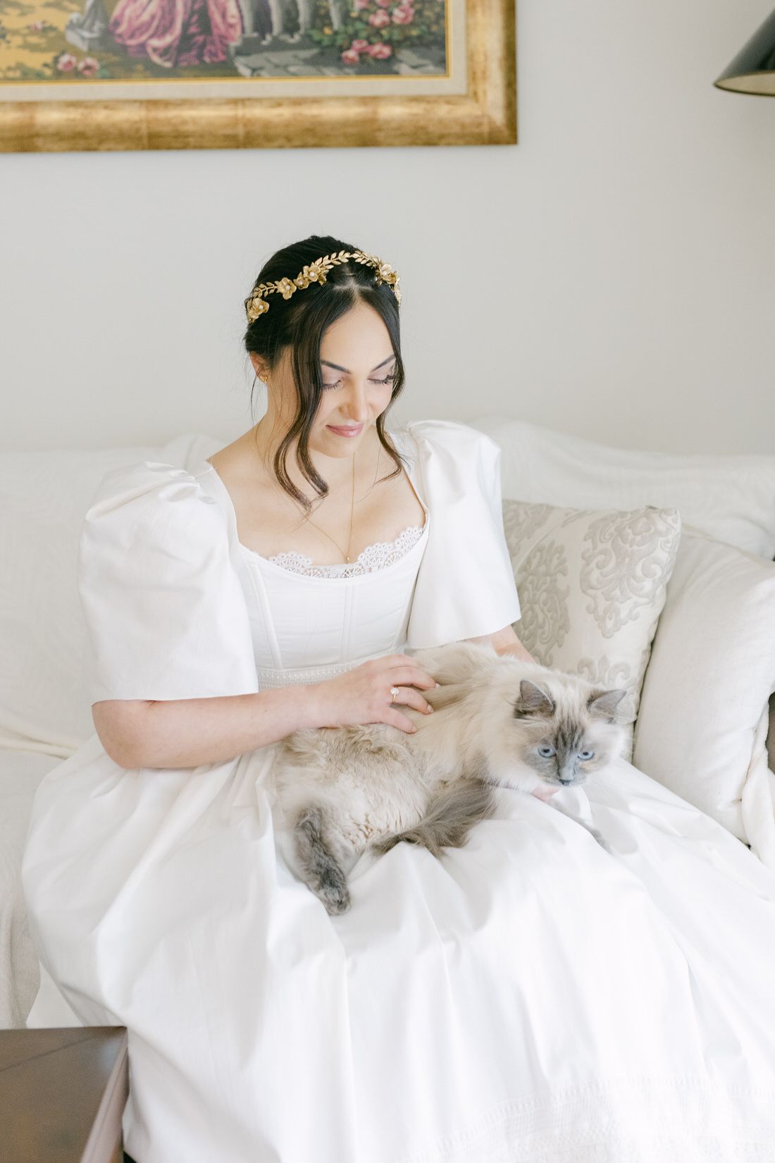 Dreamy scene of a bride in a white dress with puffy sleeves and gold crown sitting on a couch with a ragdoll cat.