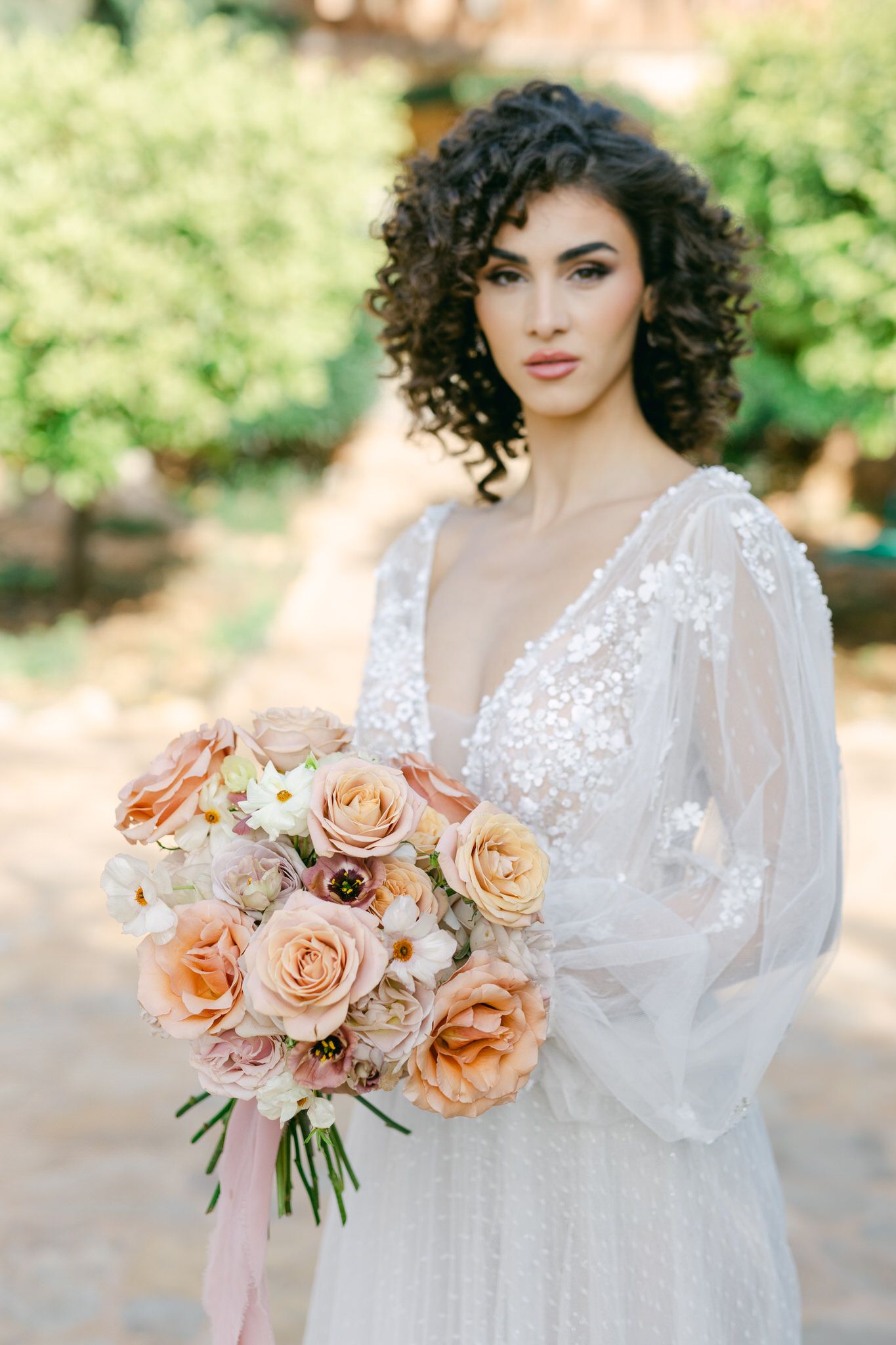 Romantic bridal portrait featuring delicate lace gown and peach-toned rose bouquet in outdoor setting.