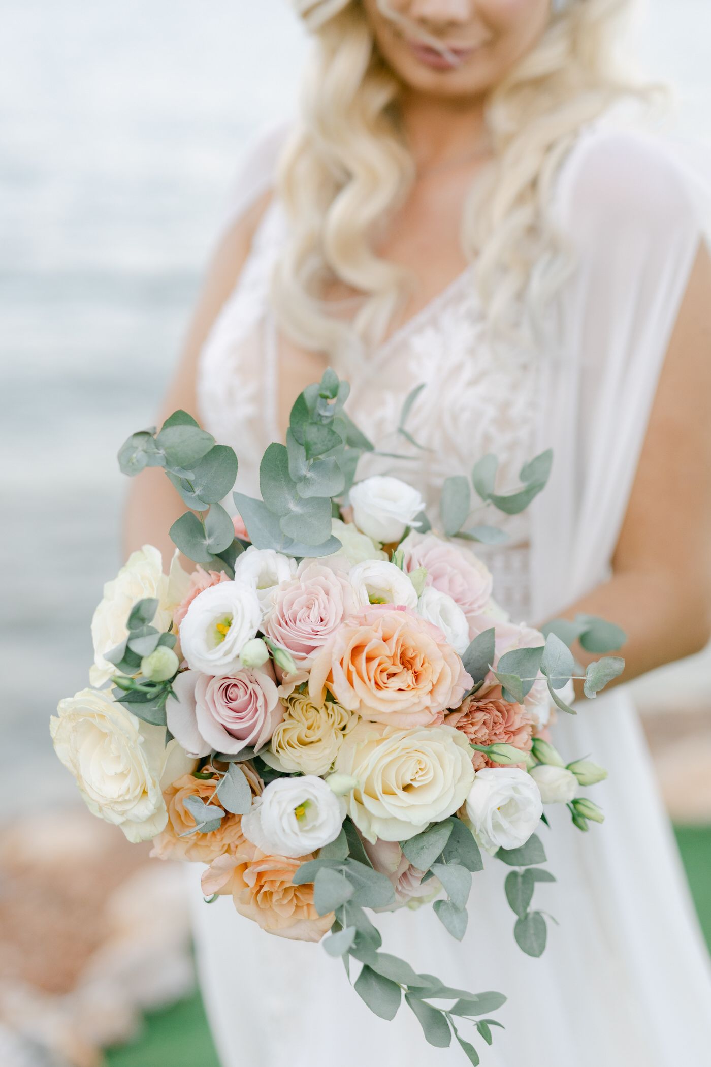A romantic bridal bouquet with blush roses, white ranunculus, peach flowers and silvery eucalyptus leaves against white dress.
