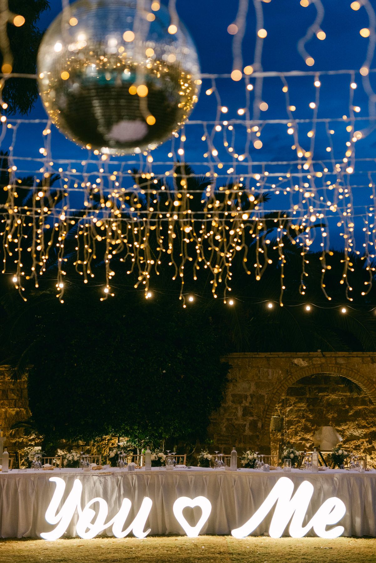 Romantic outdoor wedding reception setup with sparkling disco ball and string lights above a 'You & Me' illuminated sign.