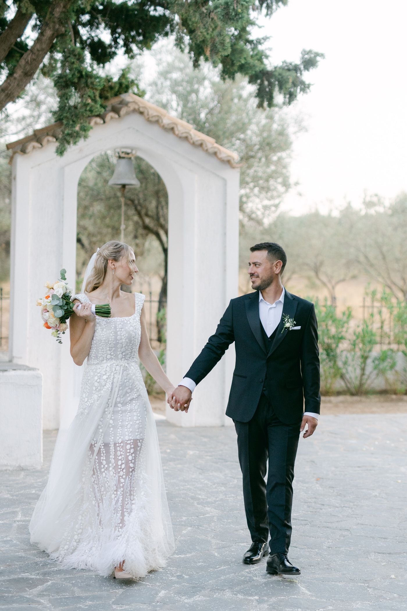 A couple in wedding attire walks hand in hand beneath a white archway surrounded by lush greenery.