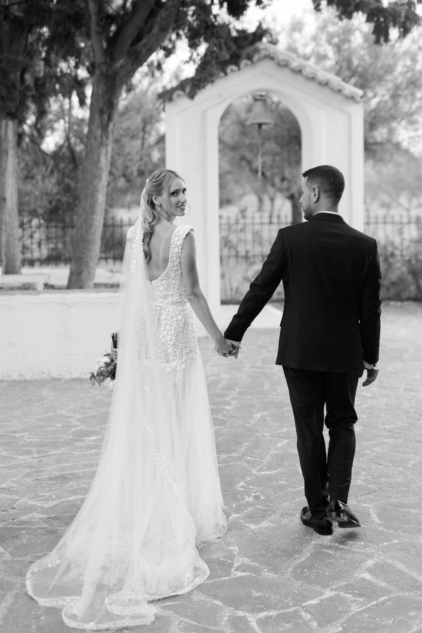 Black and white photo of a bride in lace gown and groom in suit holding hands walking toward a church archway.