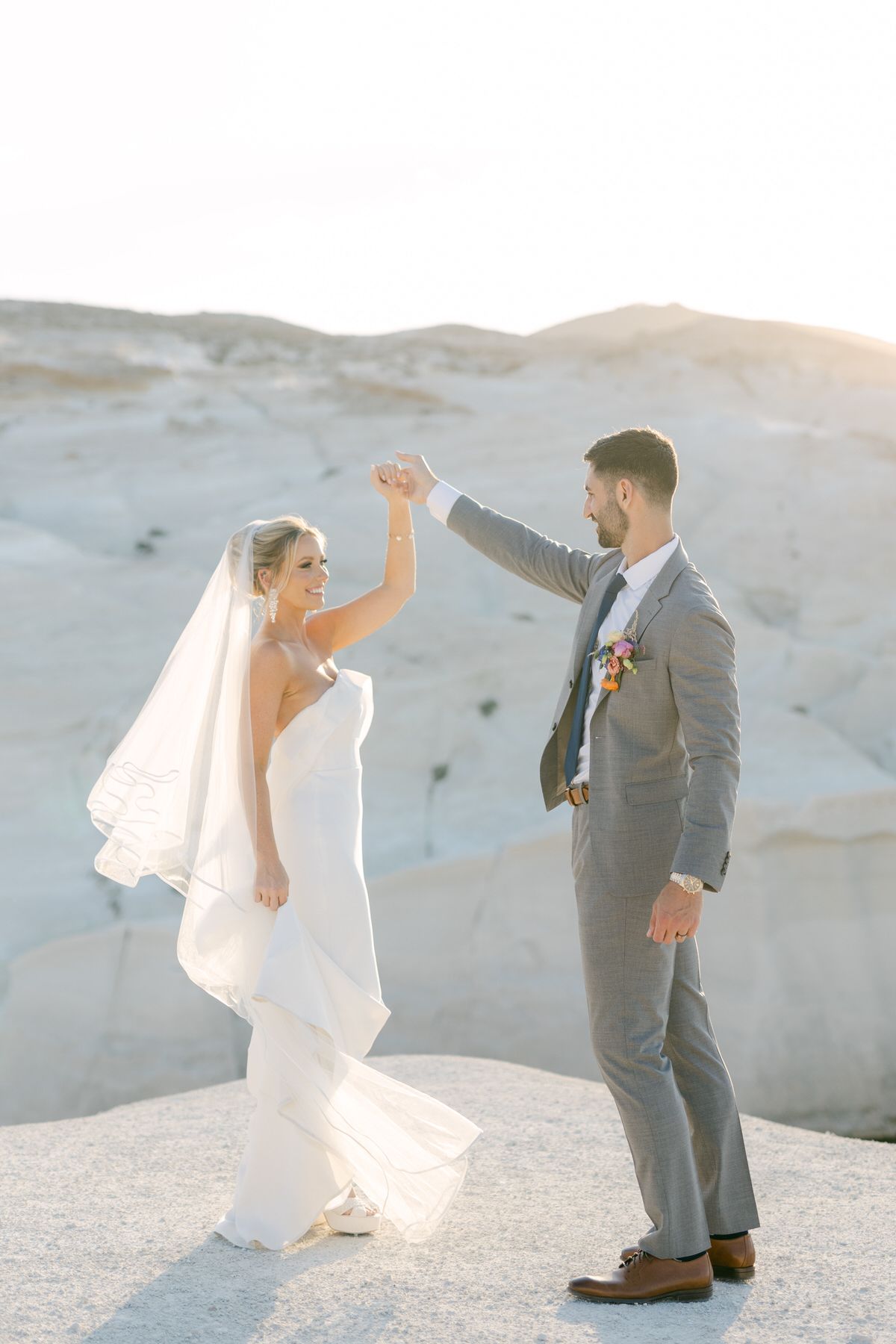 A wedding couple celebrates with raised hands against a scenic desert mountain backdrop at sunset.