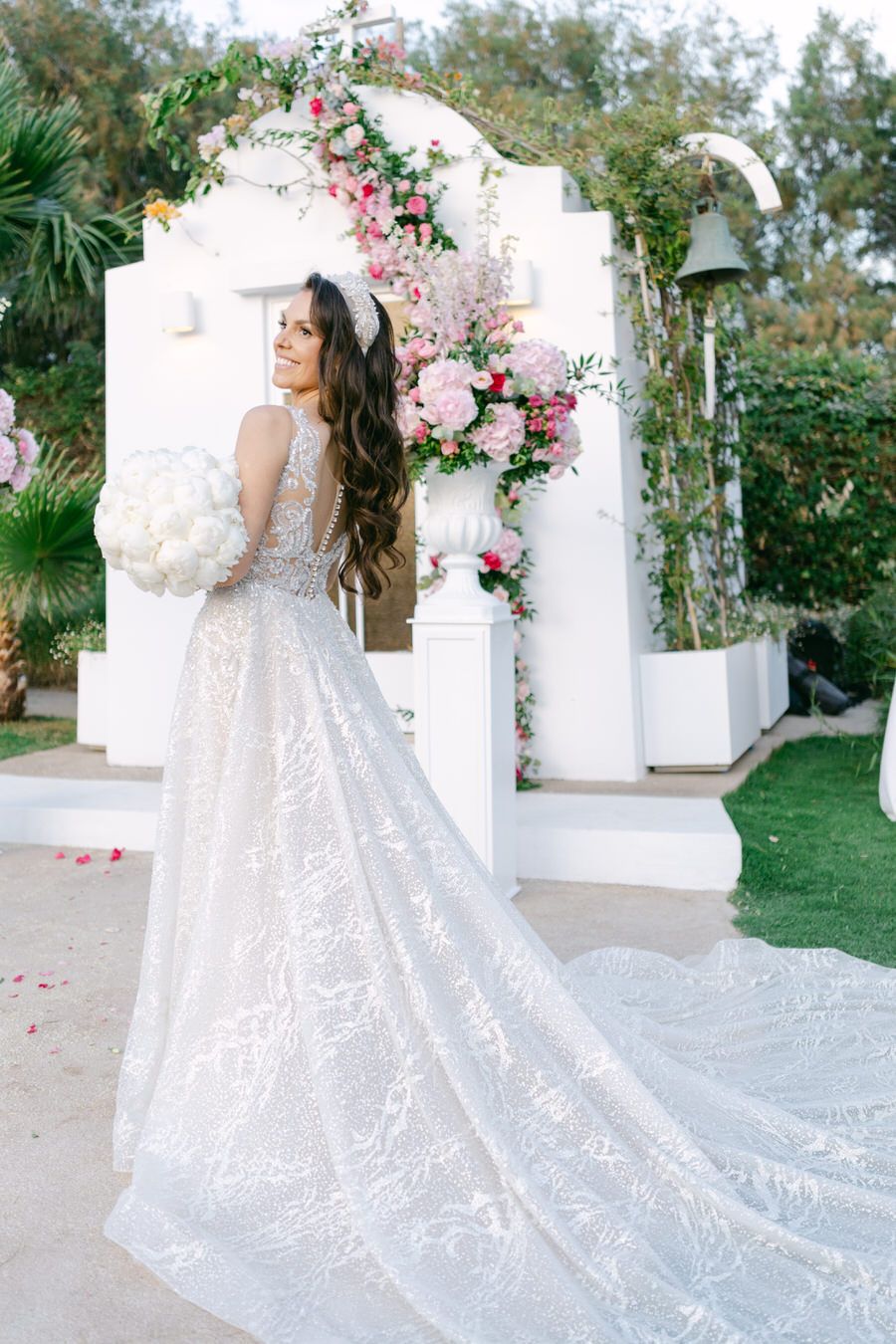 Elegant wedding dress with long train in front of floral arch decorated with pink roses.