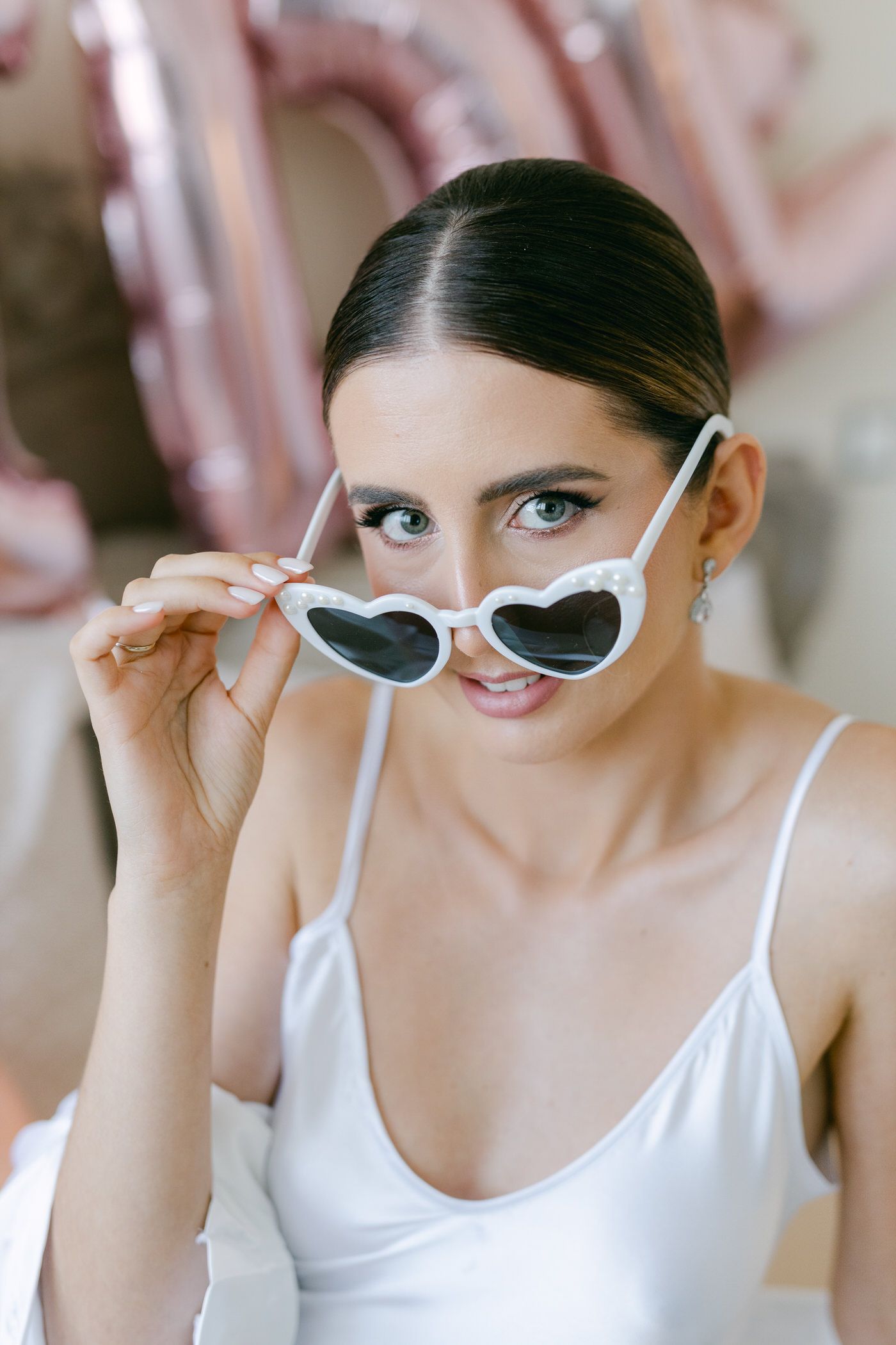 Stylish heart-shaped white sunglasses worn with a white tank top against a soft pink background.
