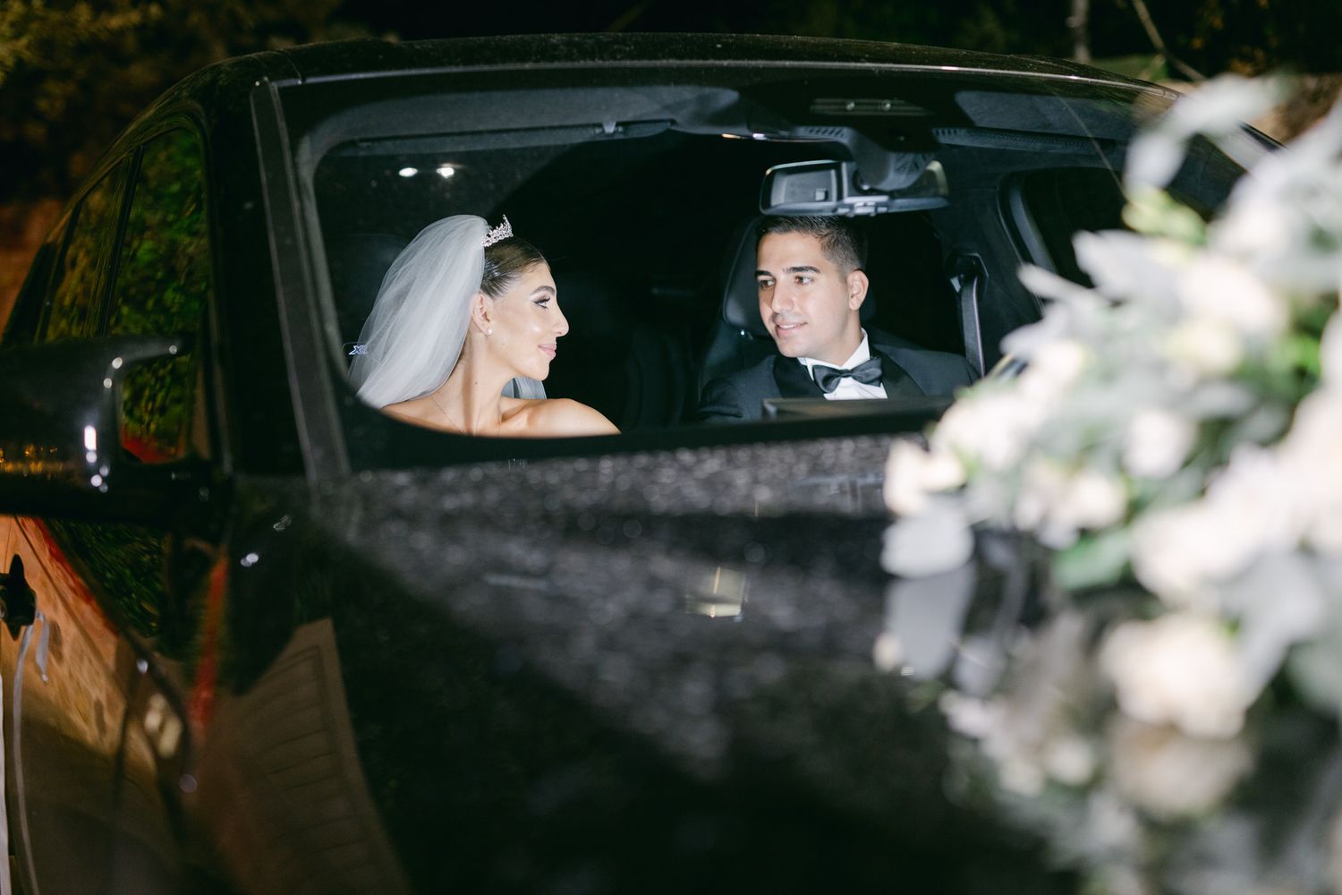 Wedding couple sitting in the backseat of a car at night with soft lights visible through the window.