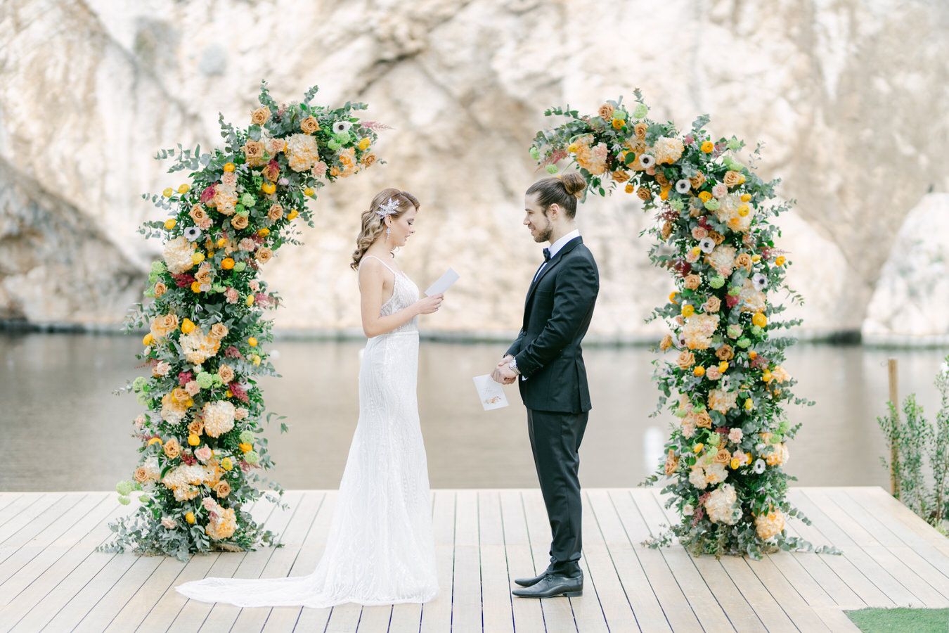 A wedding couple stands on a wooden deck by water under a floral arch with peach and white blooms.