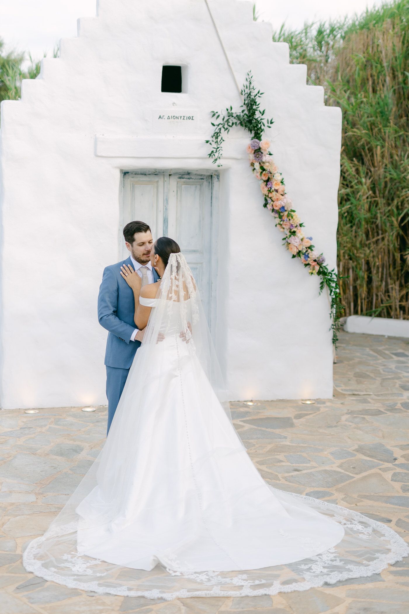 A newlywed couple embraces in front of a white stucco church entrance adorned with floral decorations.