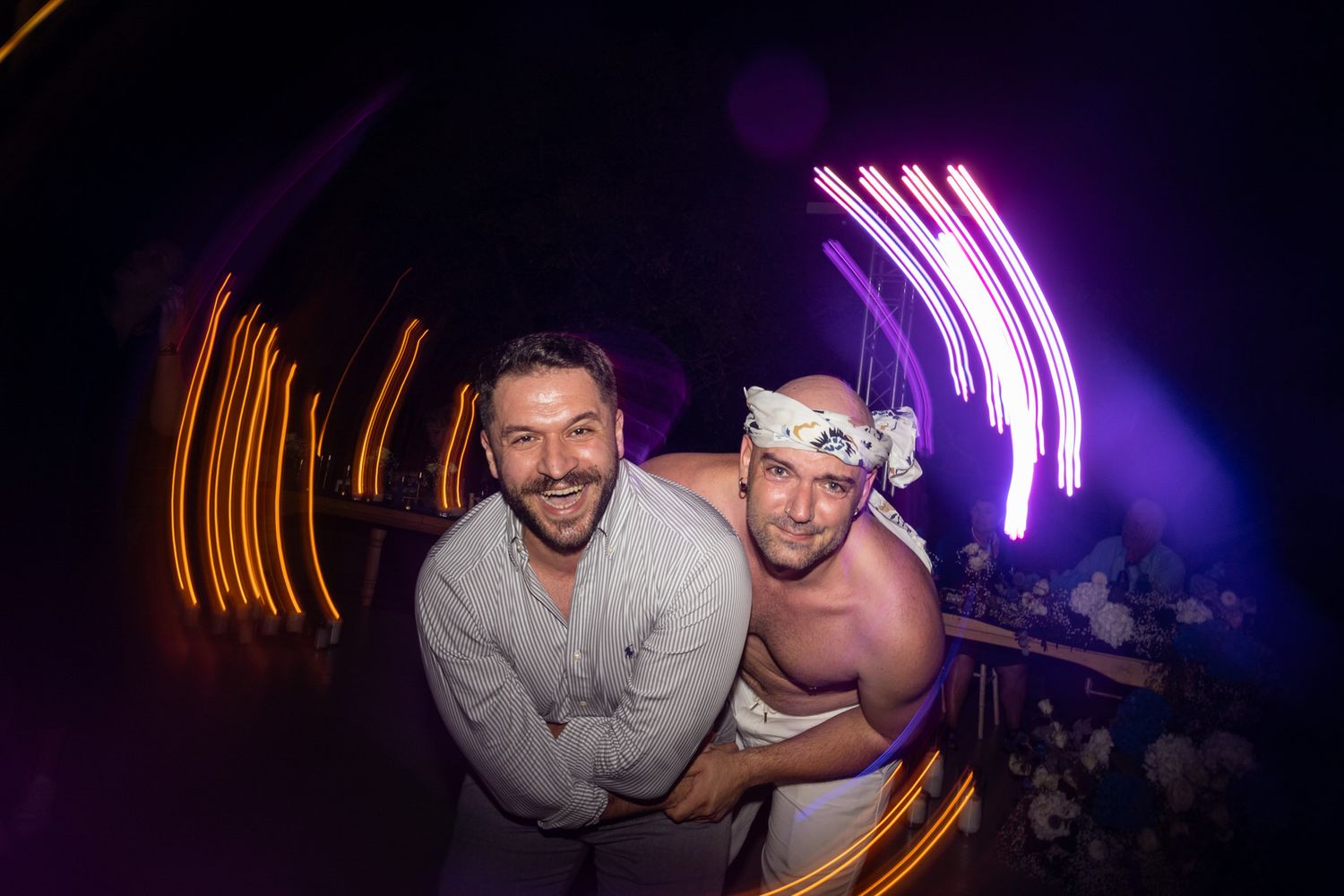 Two people pose together at a nightclub with colorful neon light streaks in the background.