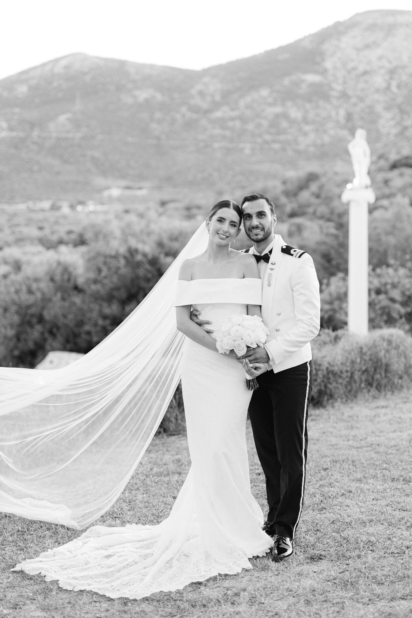 A black and white wedding photograph showing a couple outdoors with mountains in the background and a flowing veil.