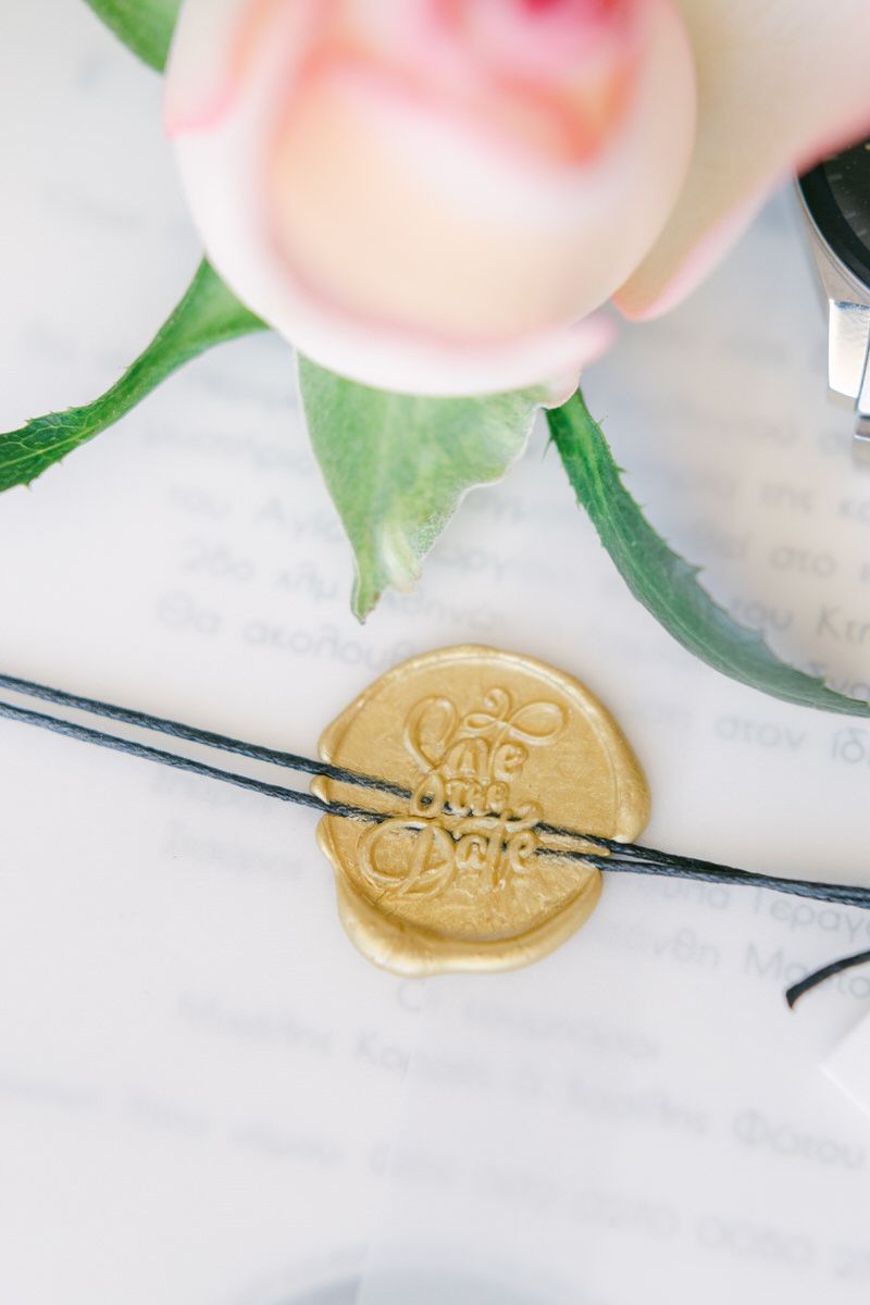 Gold wax seal with elegant script text sits beside a pink rose and green leaves on white paper.