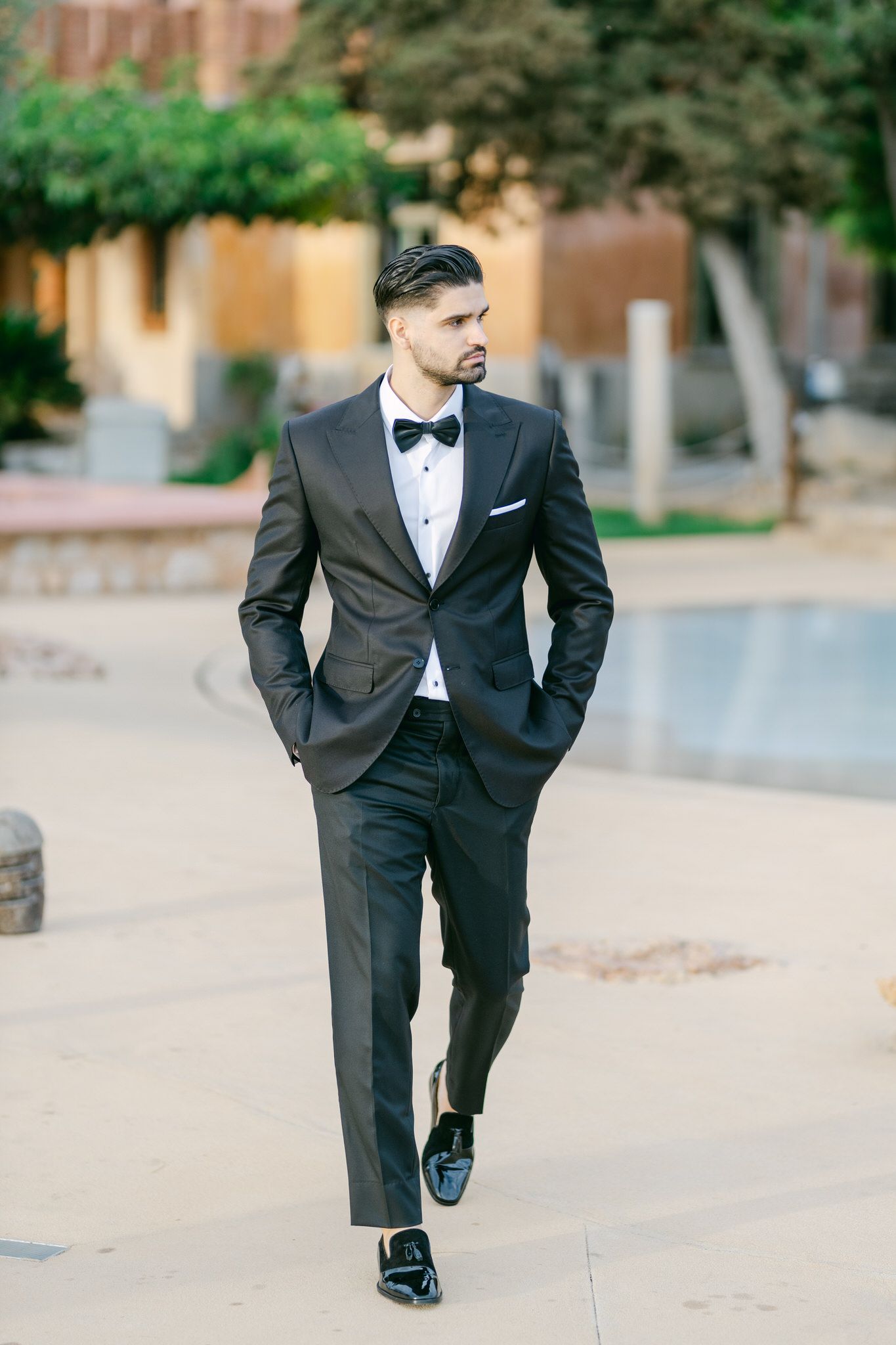 Stylish groom in classic black tuxedo and bow tie walks confidently through outdoor venue.